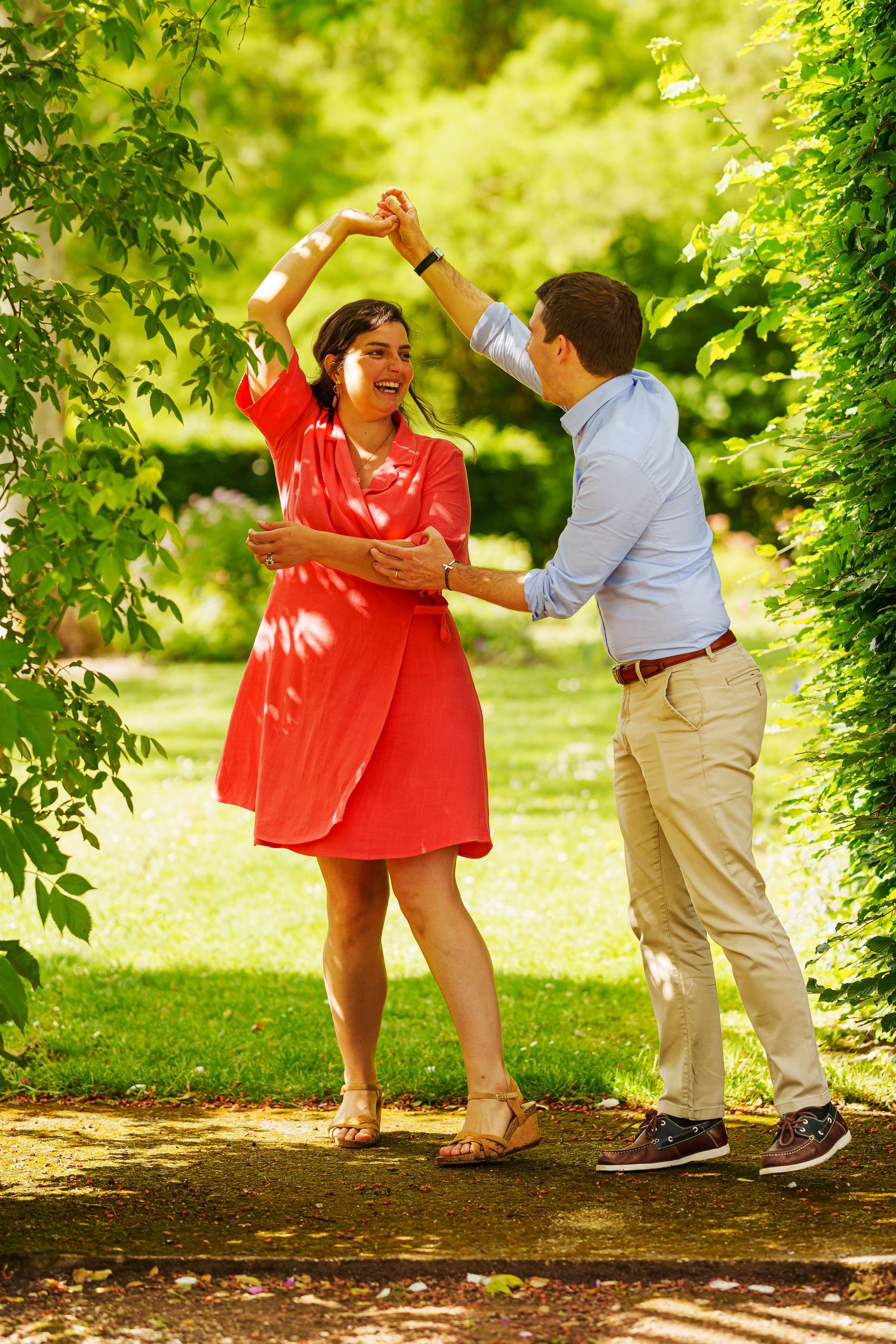 Couple dancing joyfully in a sunny green park, woman in red dress being twirled by man in blue shirt.