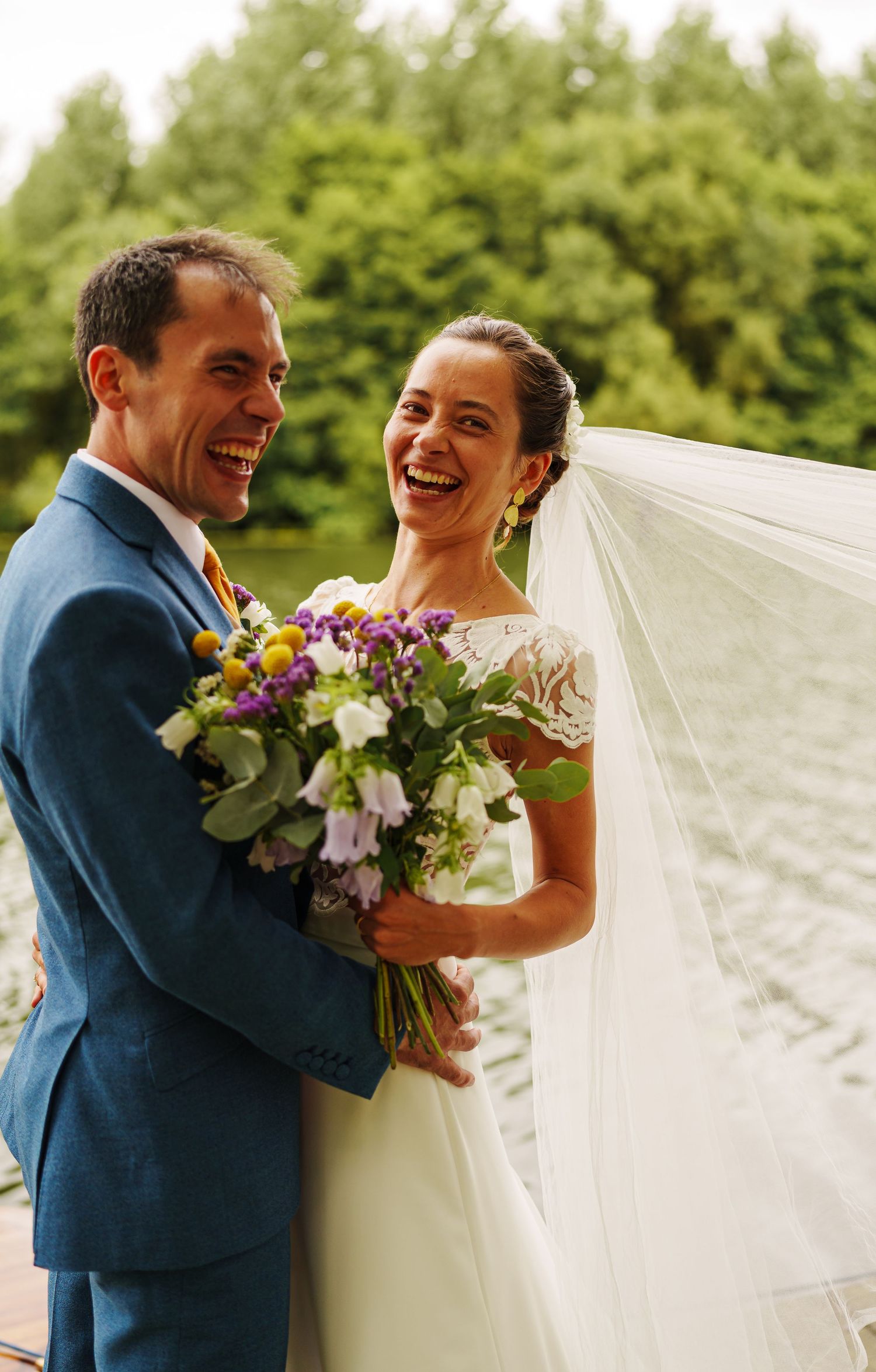 A couple laughs together while holding a colorful bouquet during their outdoor wedding celebration.
