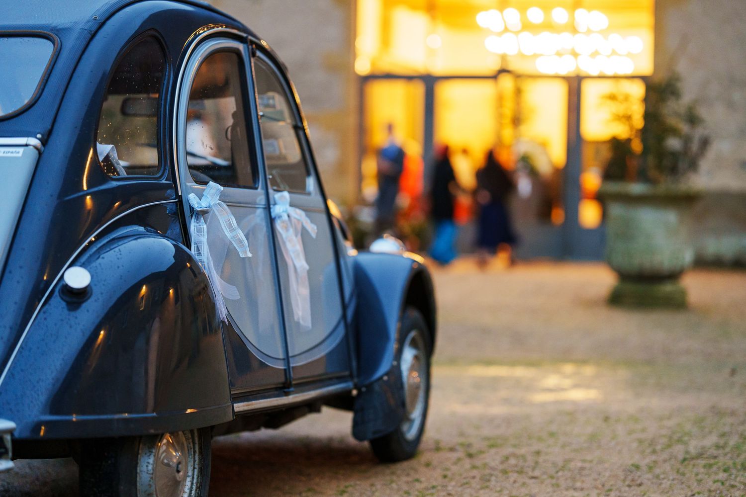 A classic blue Citroen 2CV car parked in front of a glowing storefront at dusk.