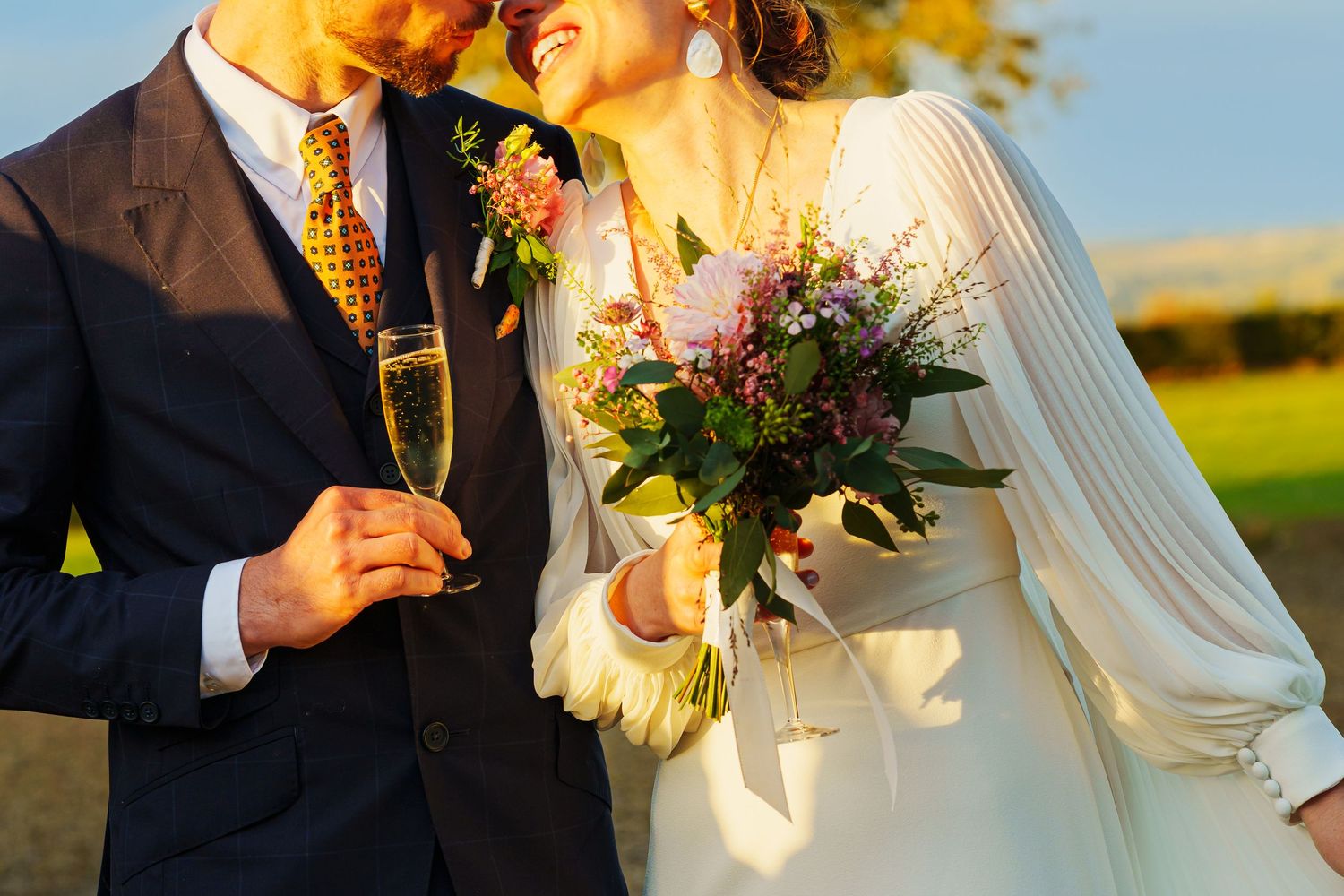 A romantic sunset wedding photo showing a couple embracing in golden light while holding a floral bouquet.