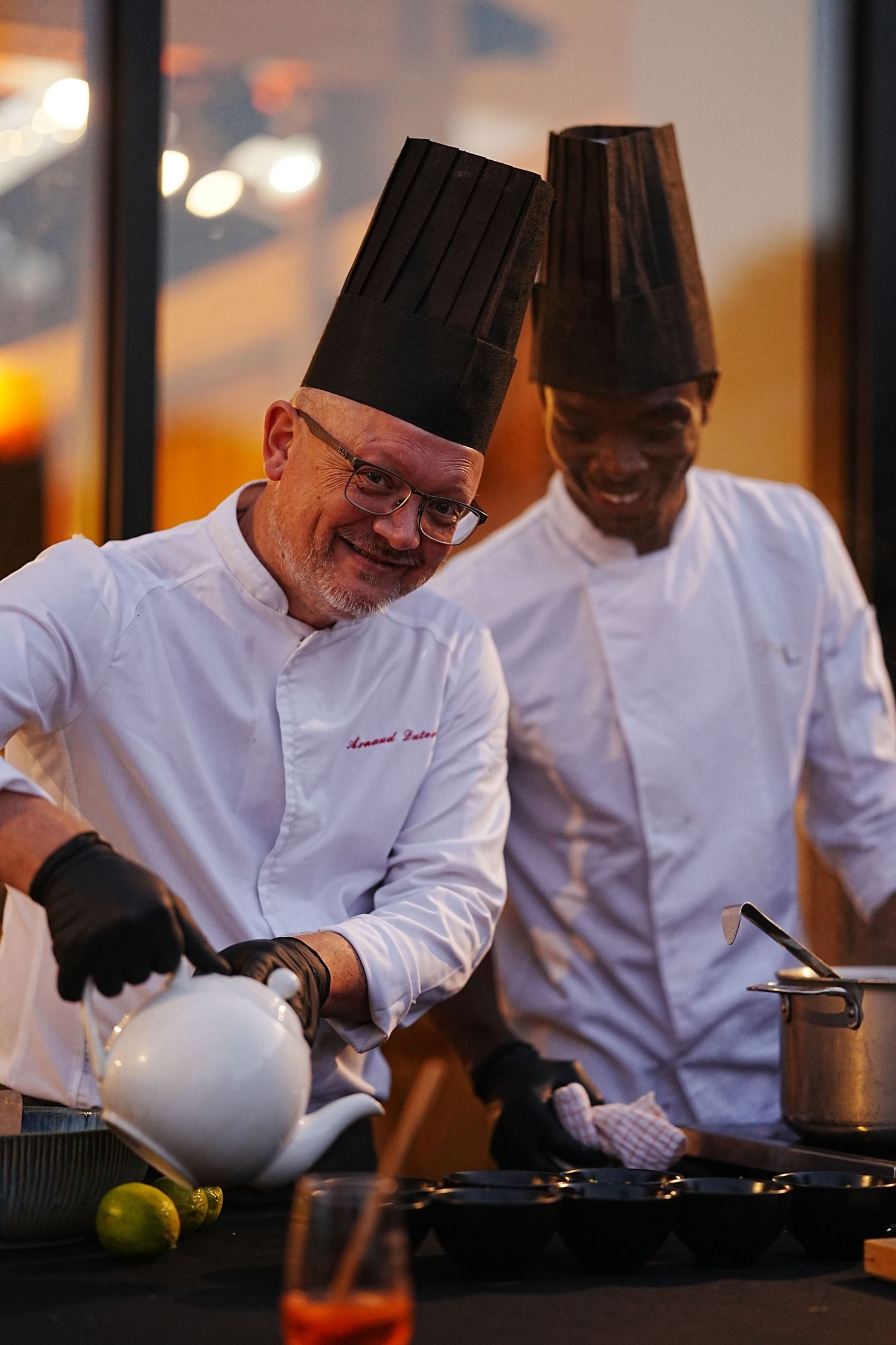 Chefs in white uniforms and black gloves prepare dishes at a restaurant counter with focused precision.