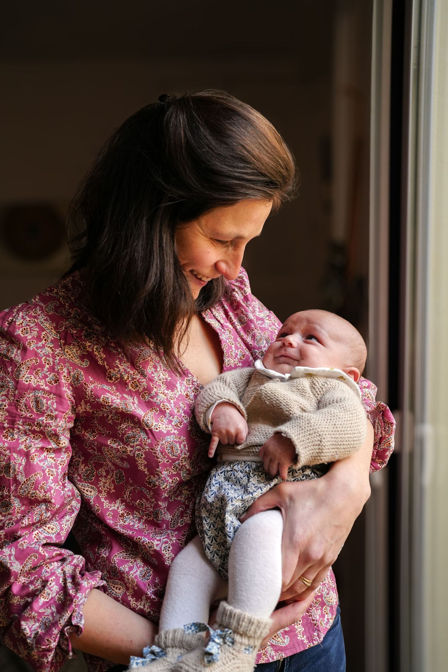 Mother in floral dress holding and feeding newborn baby by window.