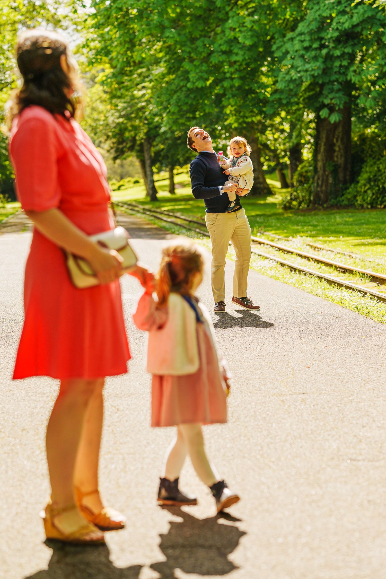 A happy family enjoys a sunny day in the park, with a man lifting a baby while a girl walks with her mother.
