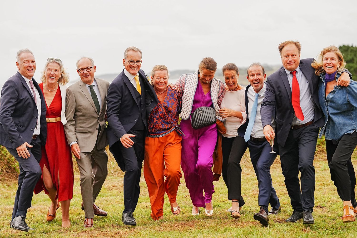 Large wedding party poses playfully in a line on grassy field during sunset.