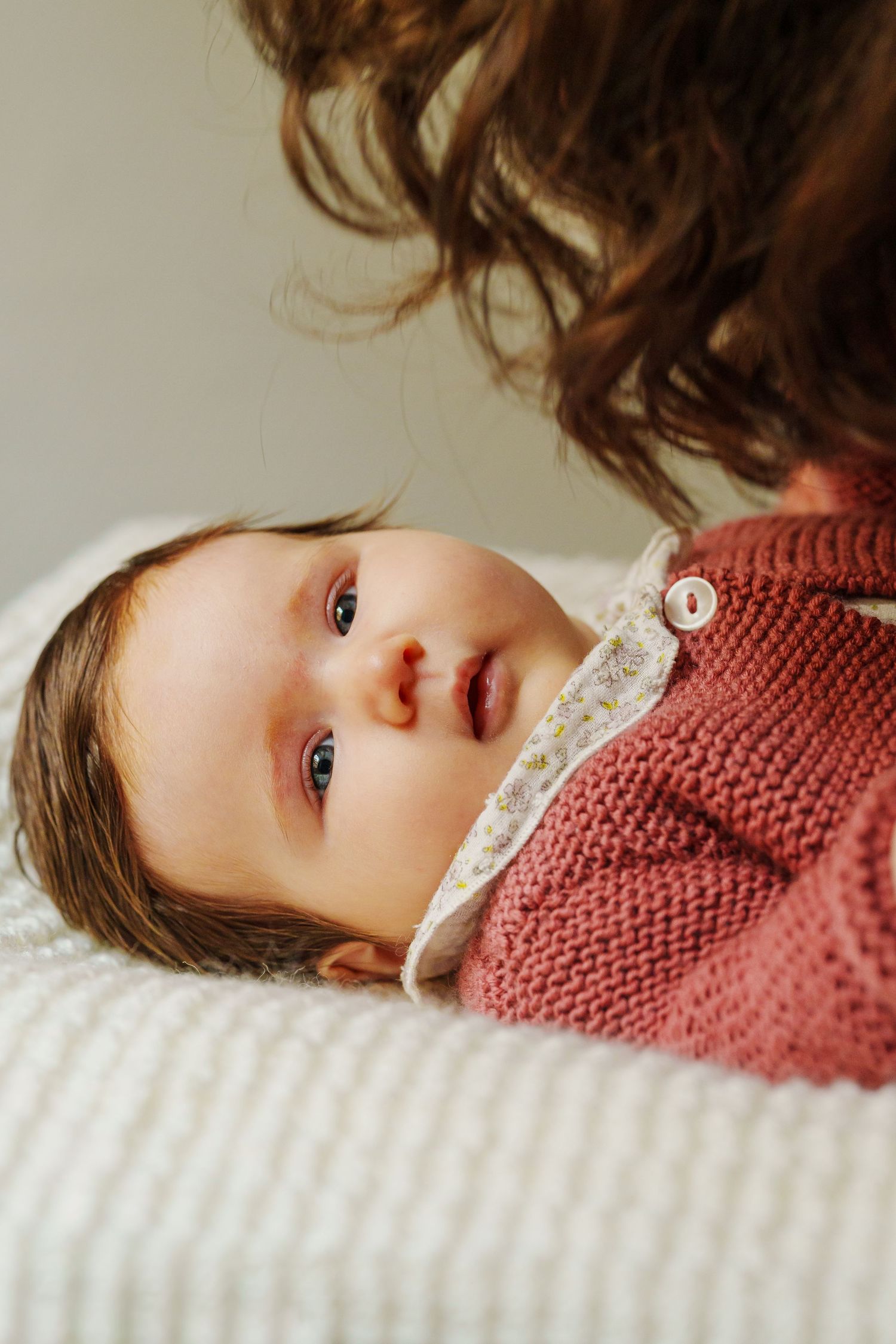 A baby in a pink knit sweater lies on a white blanket, gazing upward while a curly-haired person looks on.