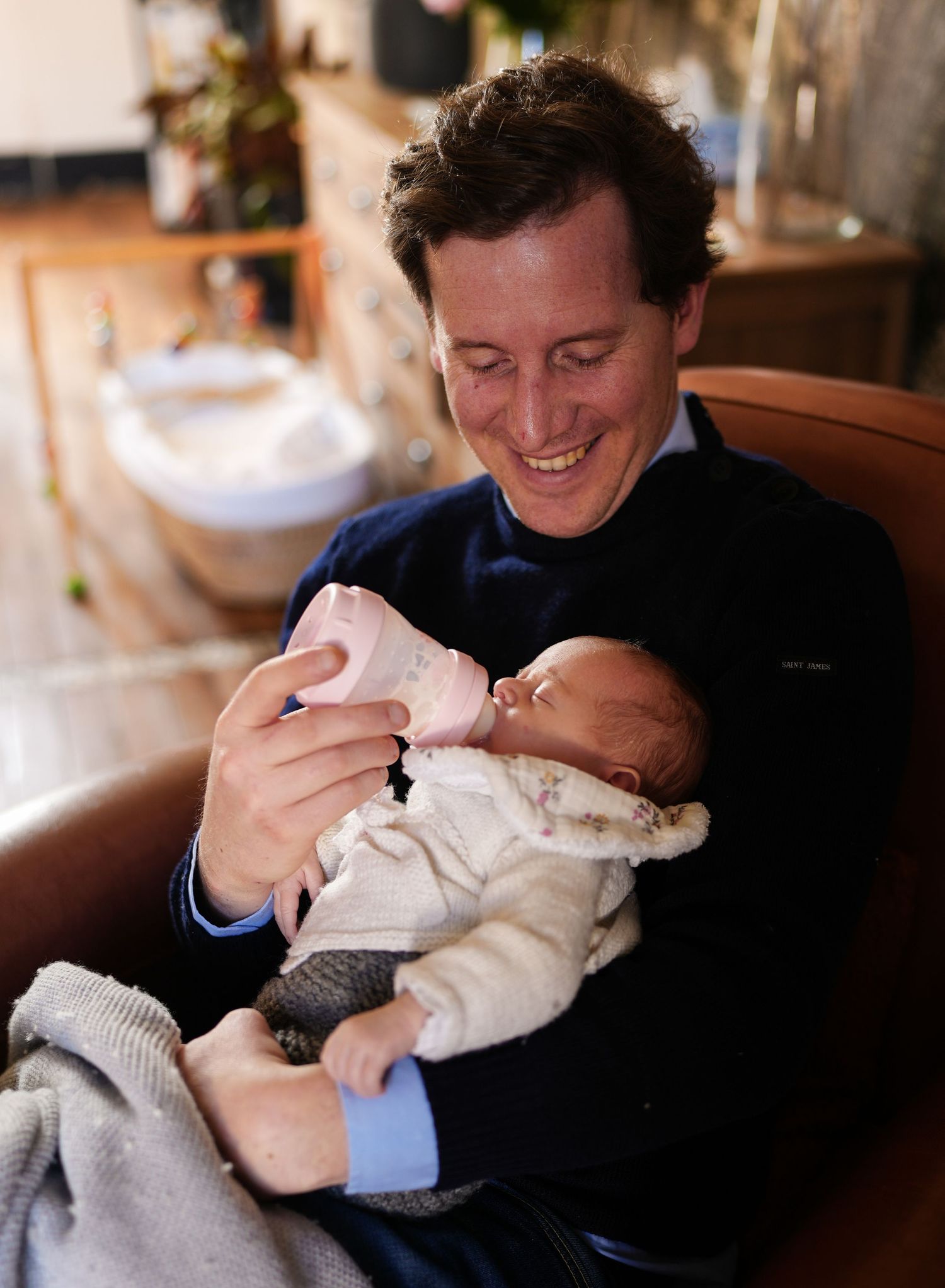Parent in navy sweater bottle feeding a newborn baby while sitting in a comfortable chair.