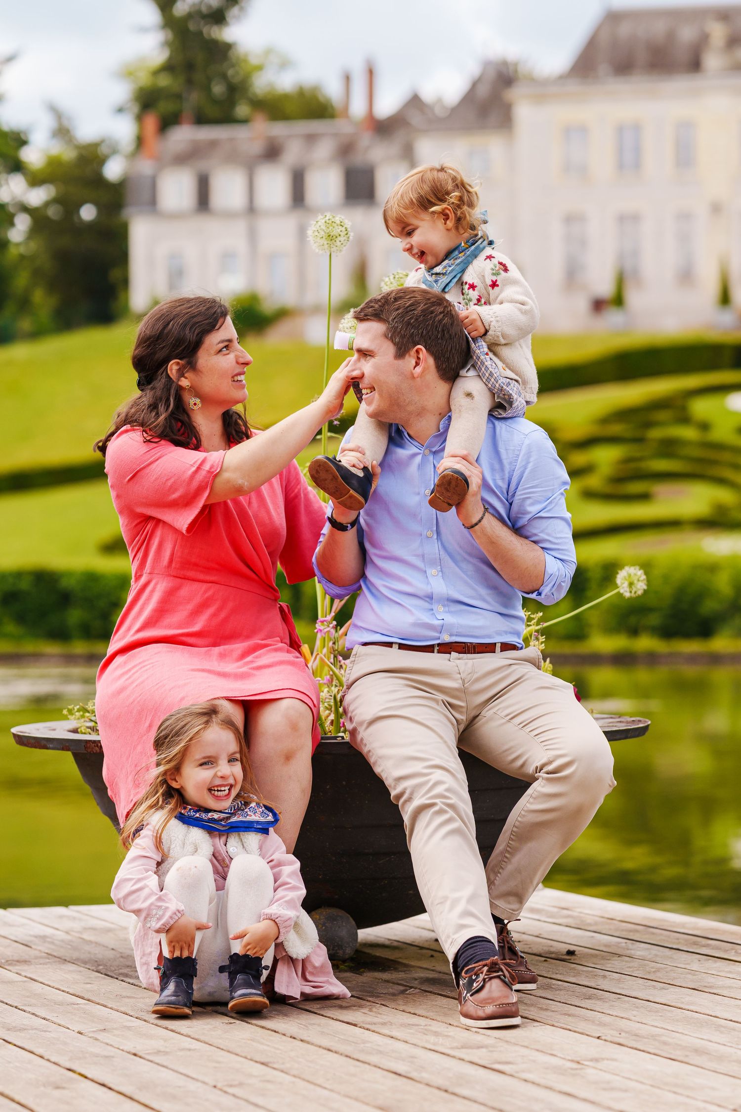 A happy family of four enjoys a sunny day outdoors, sitting near a pond with a chateau in the background.
