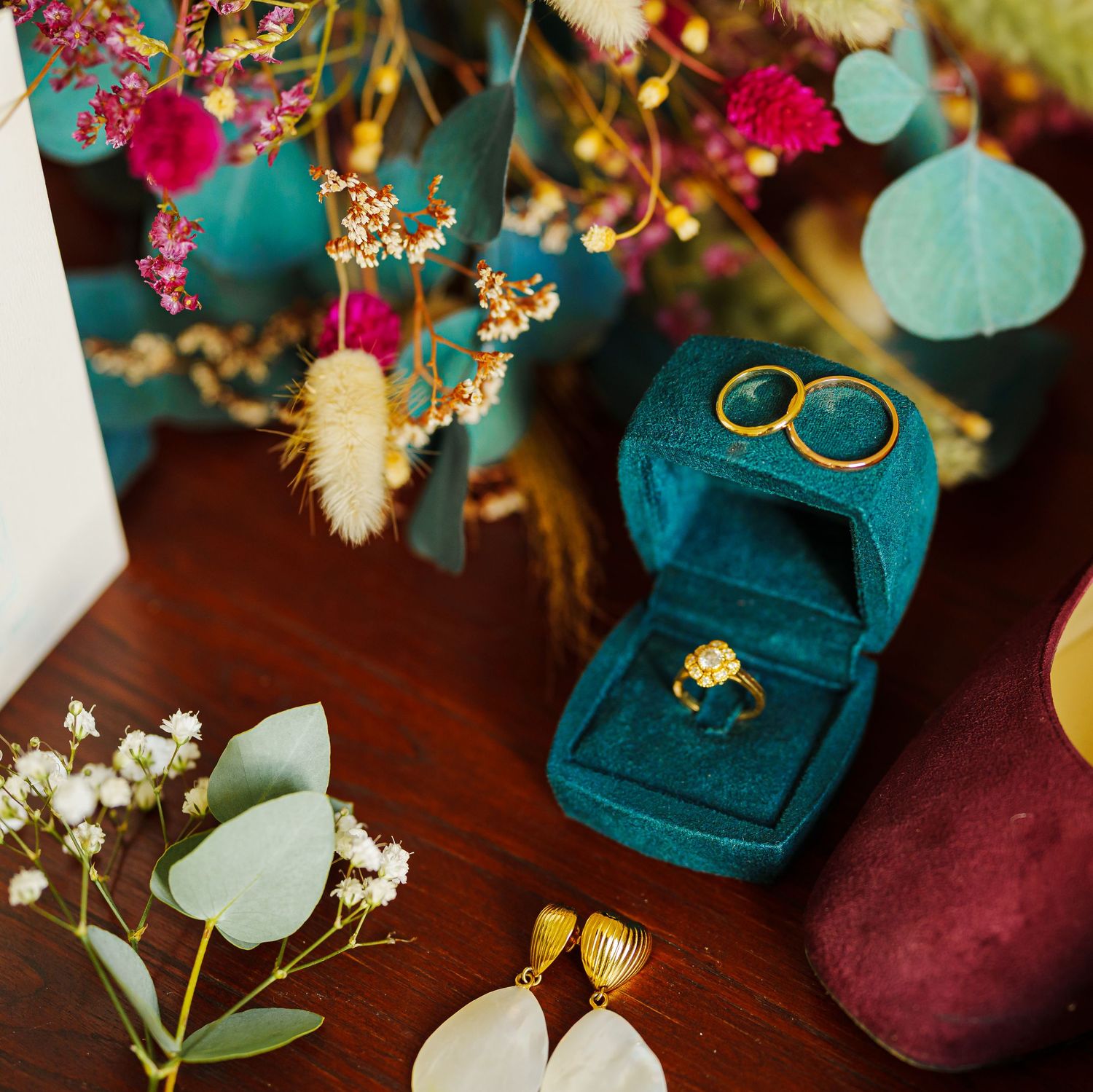 Wedding details with teal velvet ring box, burgundy accessories, delicate flowers and greenery on dark wooden surface.