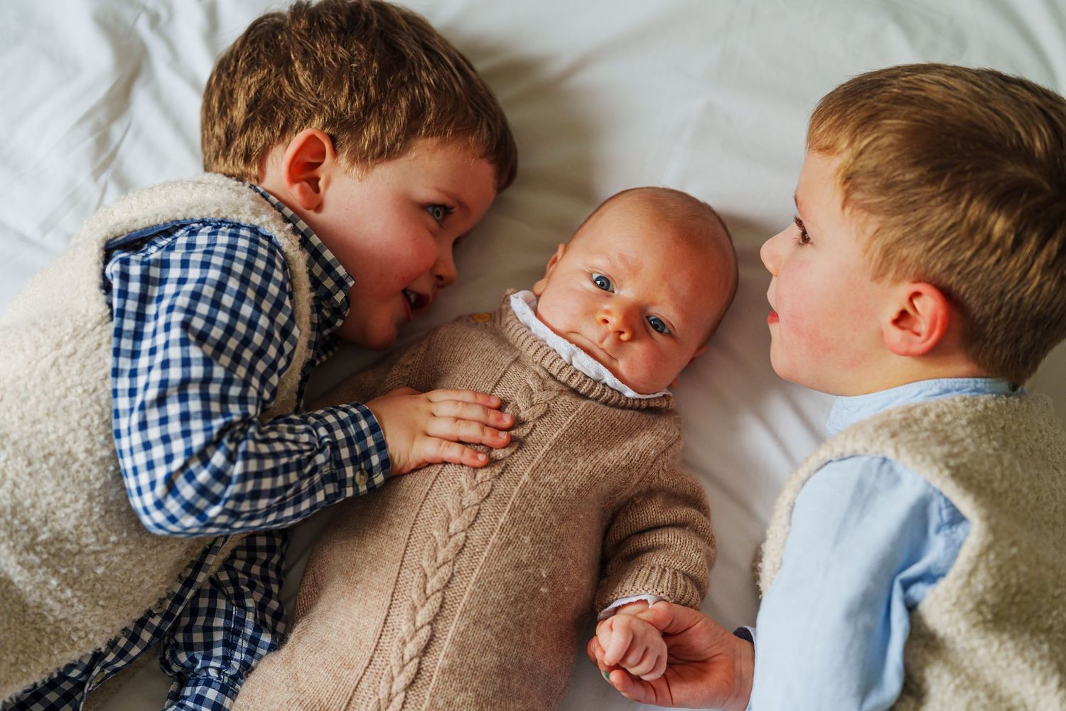 Two young boys lying on a white bed with a newborn baby sibling between them, wearing knit outfits.