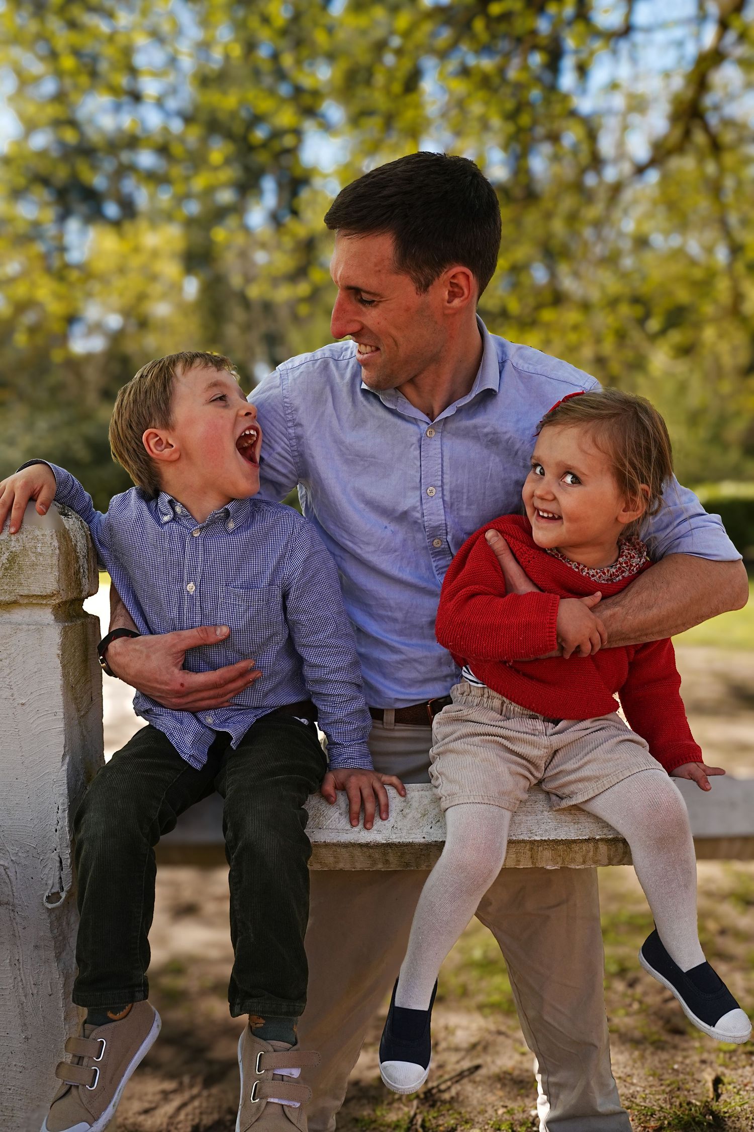 Family sitting together on wooden fence sharing laughs in sunlit park setting.