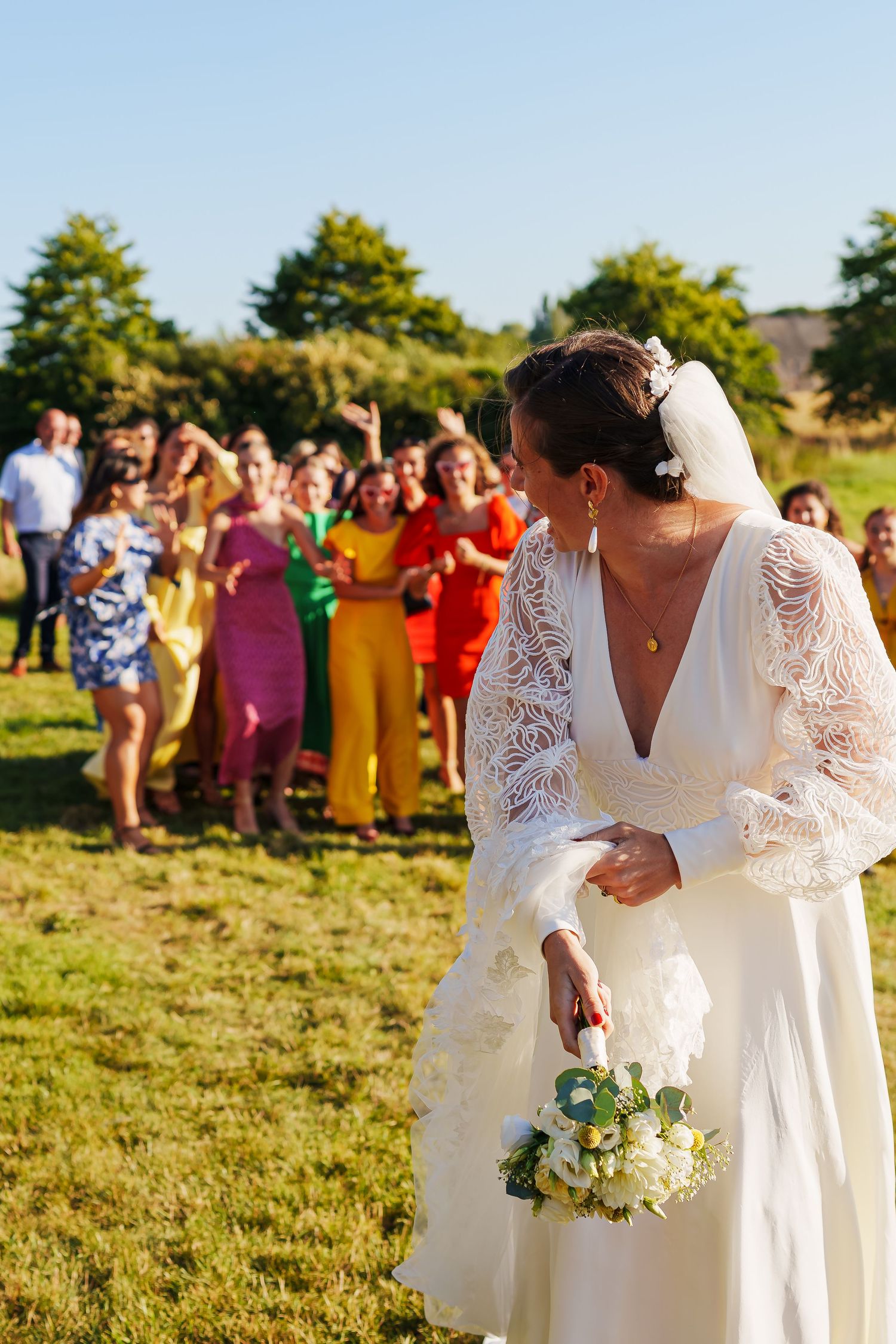 Wedding couple in white dresses celebrate with guests in colorful attire on sunny field.