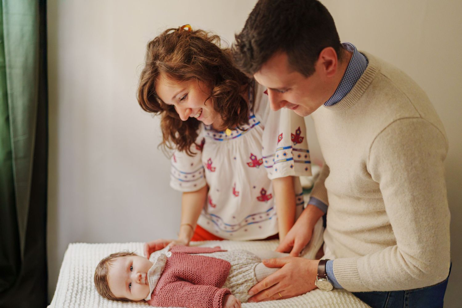 Parents smiling lovingly at their newborn baby lying on a changing table in a bright, cozy nursery room.