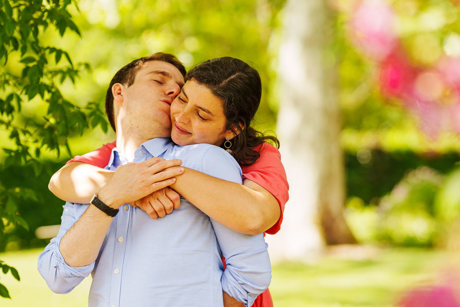 Couple embracing and kissing outdoors in a sunny green park surrounded by blooming trees.
