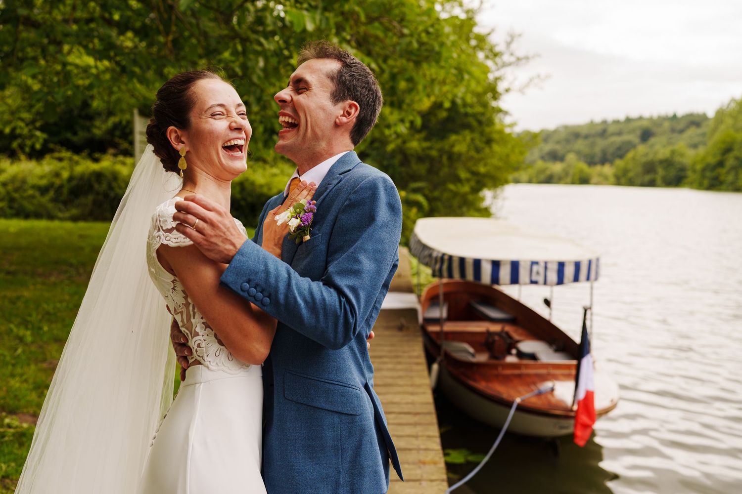 Newlyweds share a joyful laugh together on a wooden dock by a lake with boats in the background.