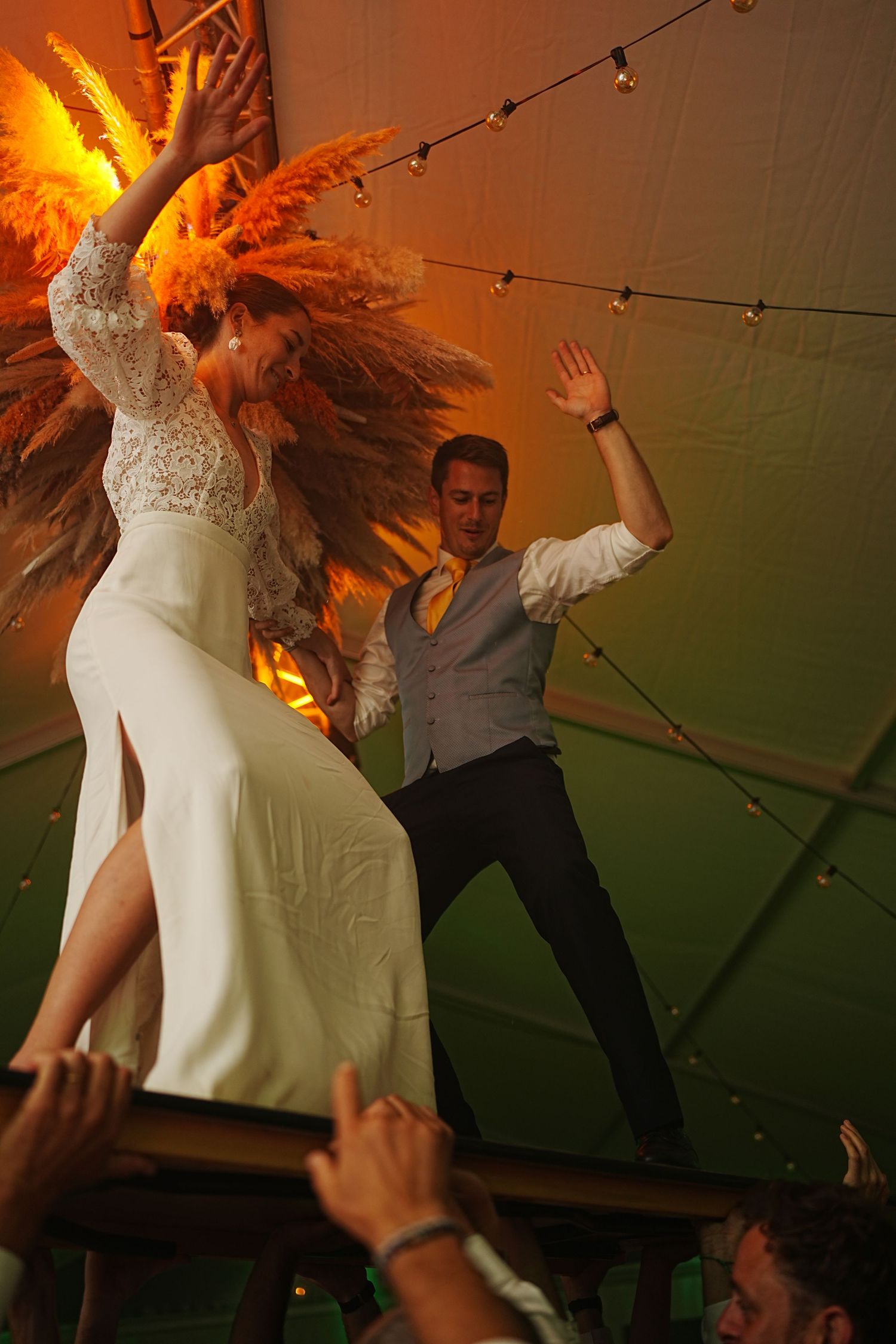 Energetic dance party scene at wedding reception with orange ambient lighting and string lights.