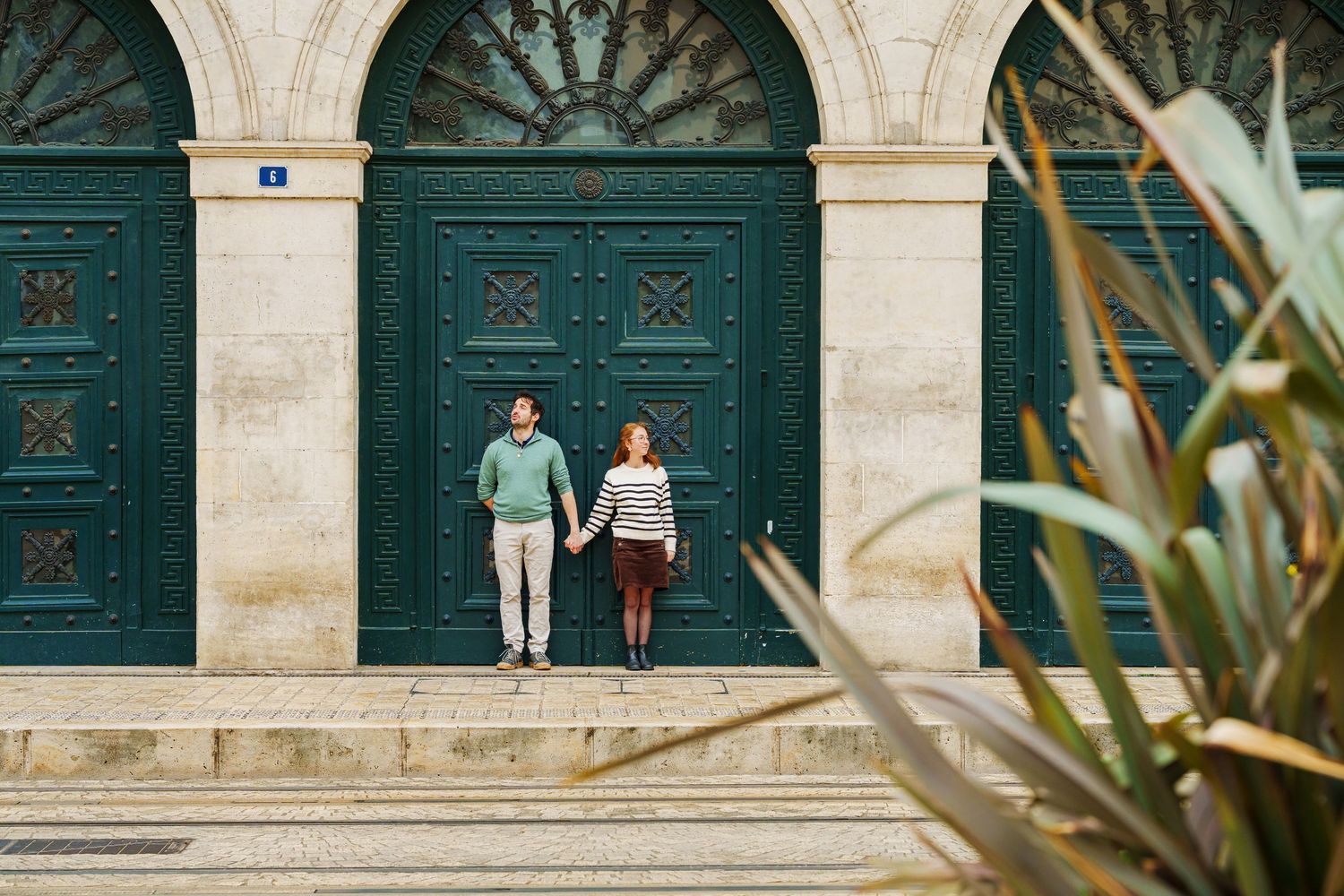 Couple holding hands on steps before large teal doors of a historic stone building.