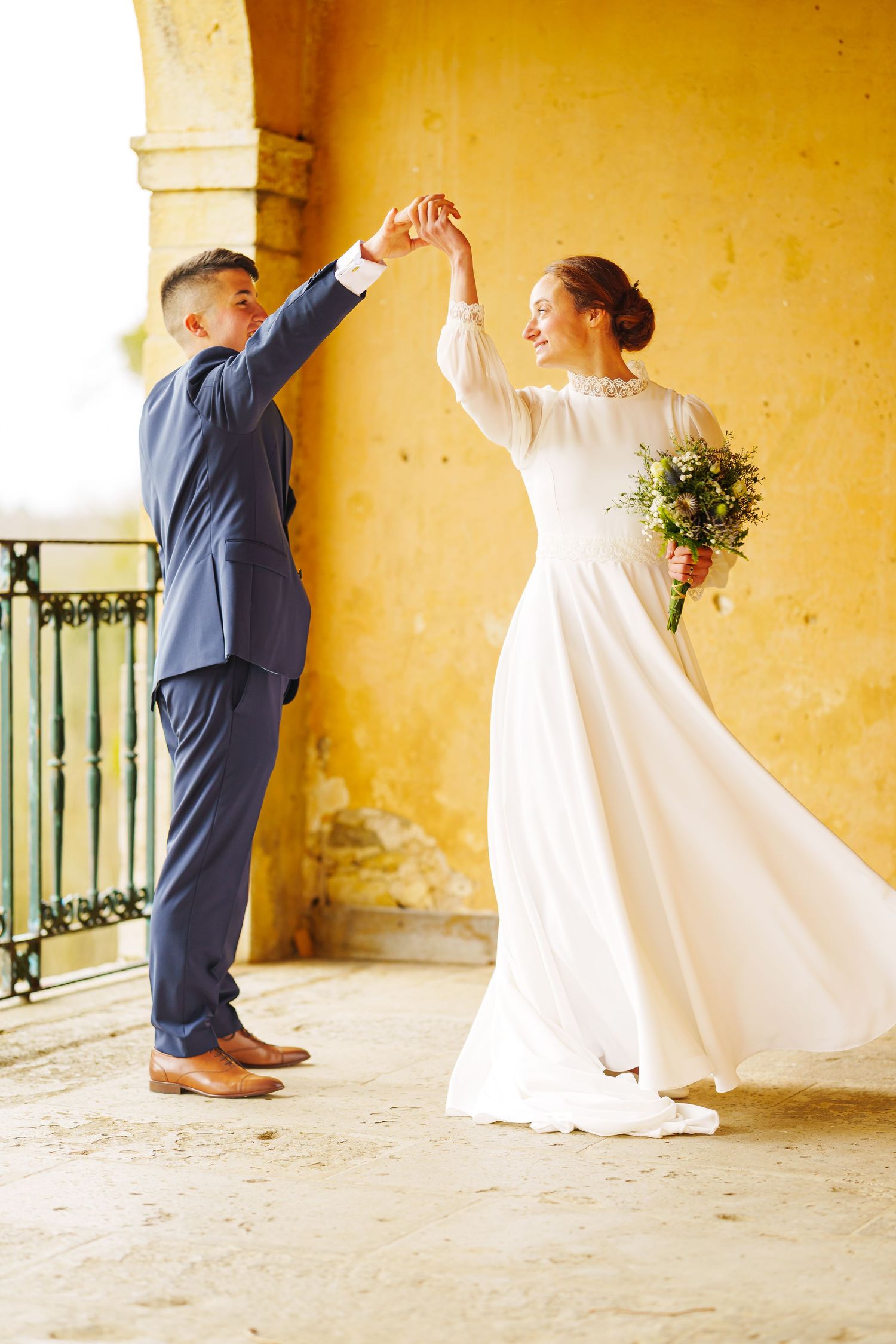 A couple in wedding attire dancing together on a terrace with yellow walls and an ornate railing.