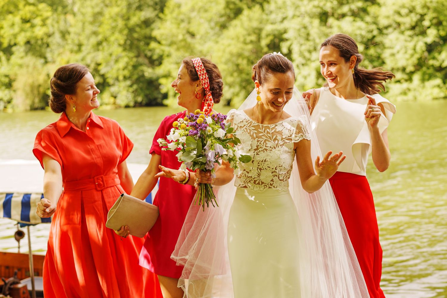 Wedding party in red and white dresses laughing together by a lake.