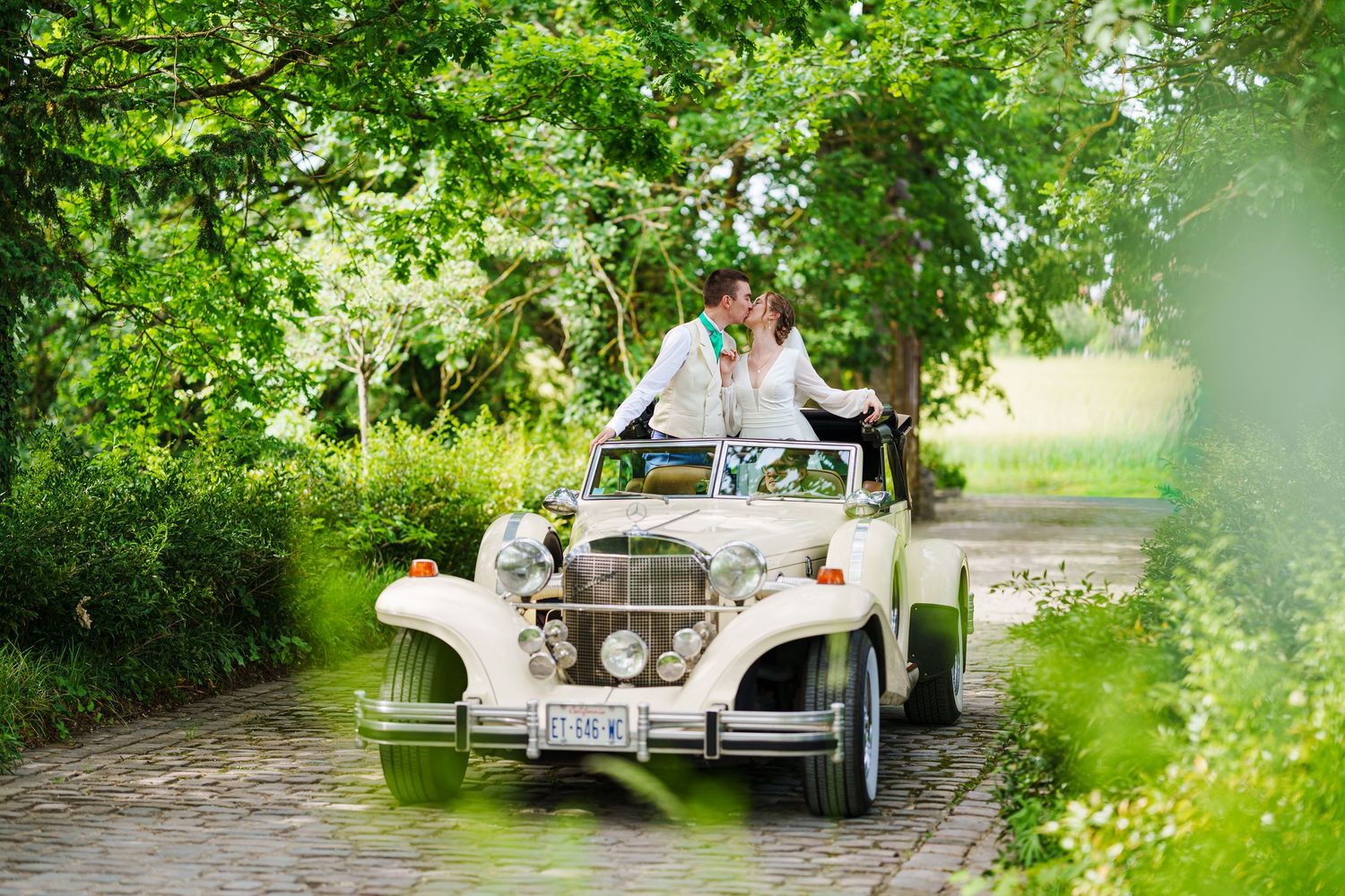 Bride and groom kissing in a vintage white convertible car on a cobblestone path surrounded by lush green trees.