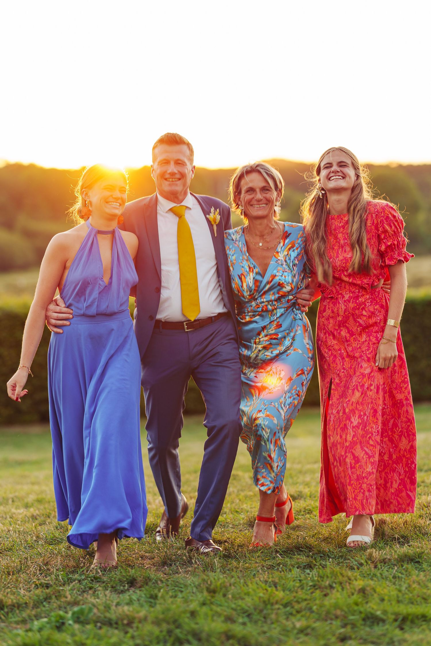Group poses together on grassy hill during golden sunset wearing formal attire in blue, red and navy colors.