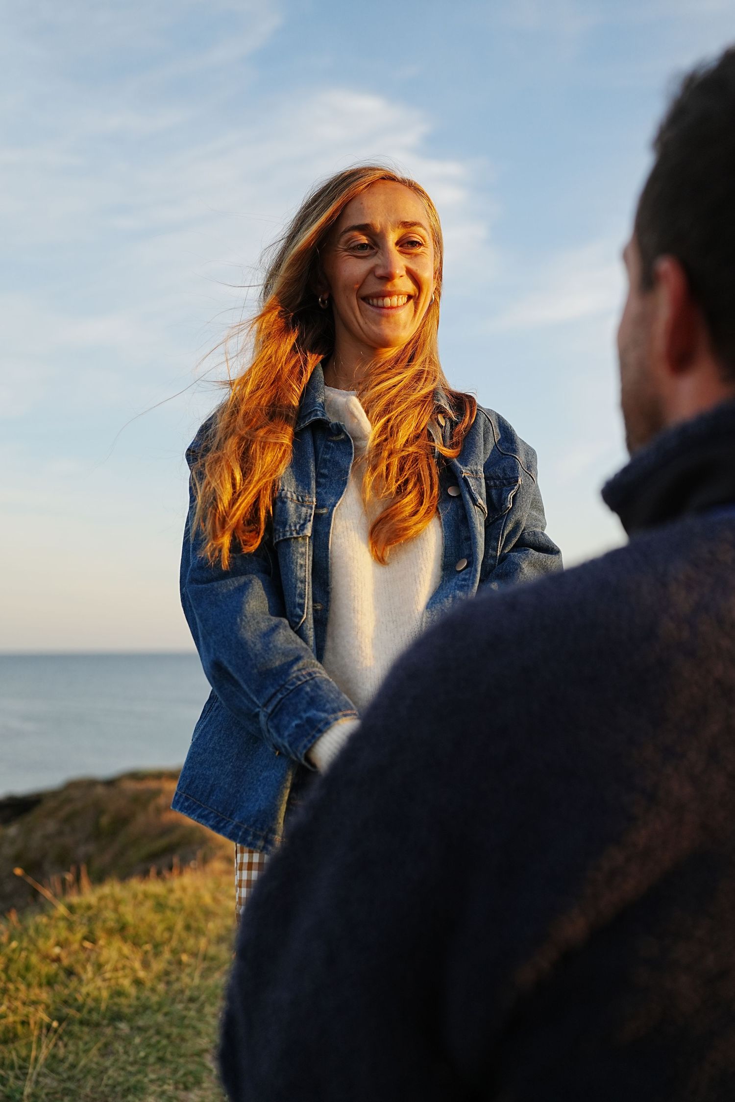 Smiling woman with red hair in denim jacket standing outdoors by the sea on a sunny day.