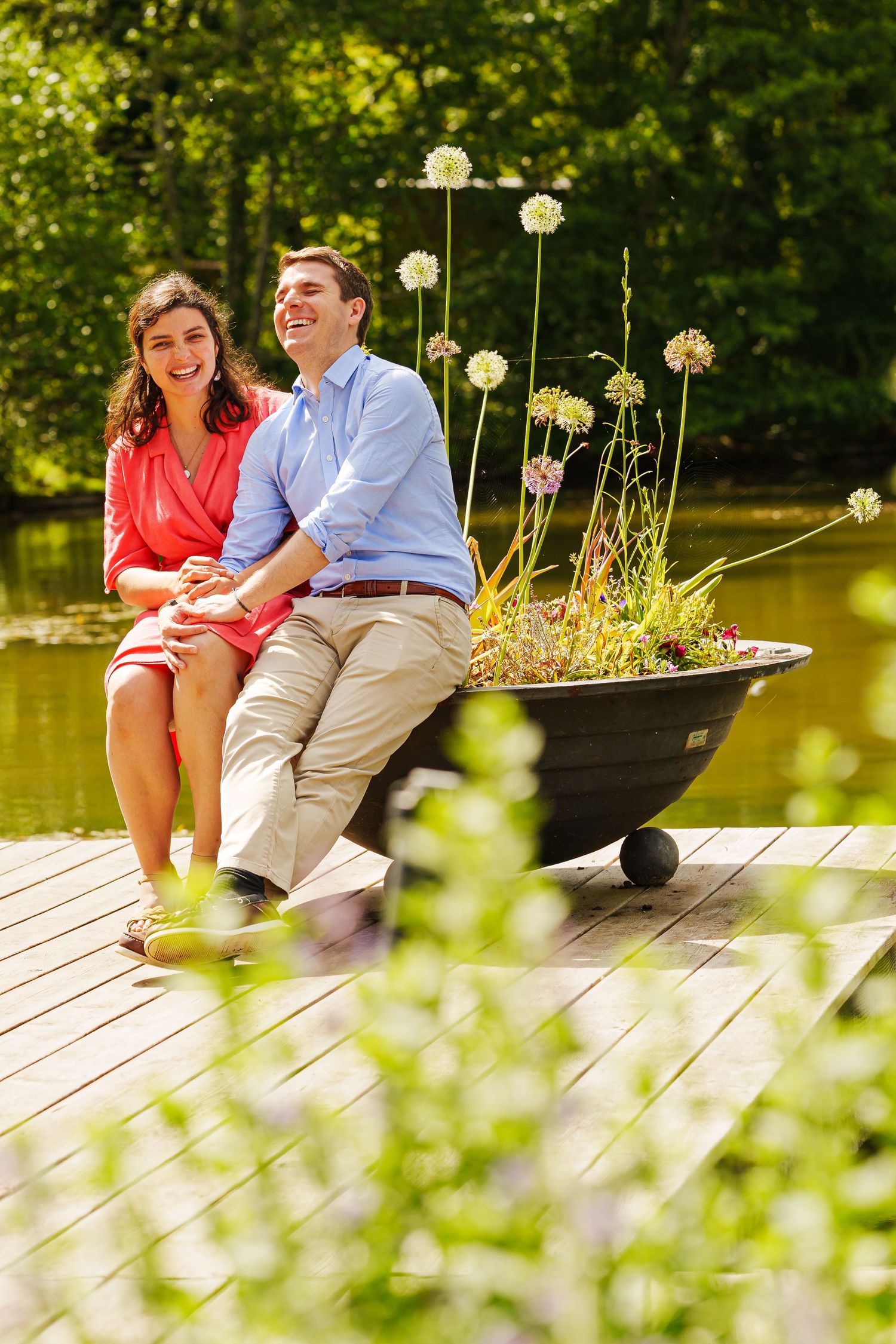 Happy couple sitting on a sunny wooden dock by a lake, surrounded by lush greenery and a planter with white flowers.