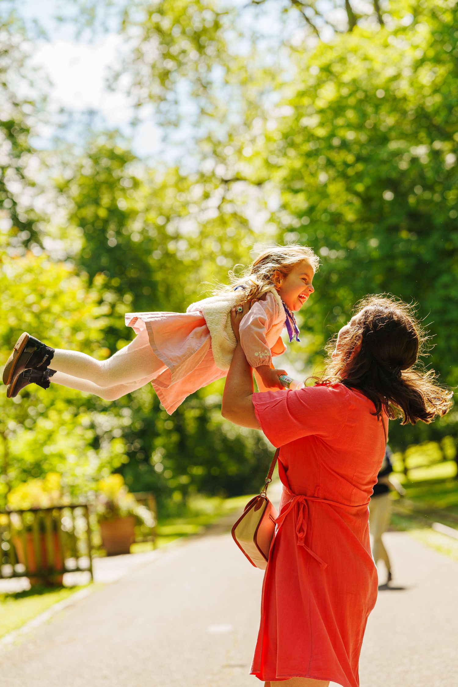 A mother in a red dress joyfully lifts her laughing daughter in a sunny green park.
