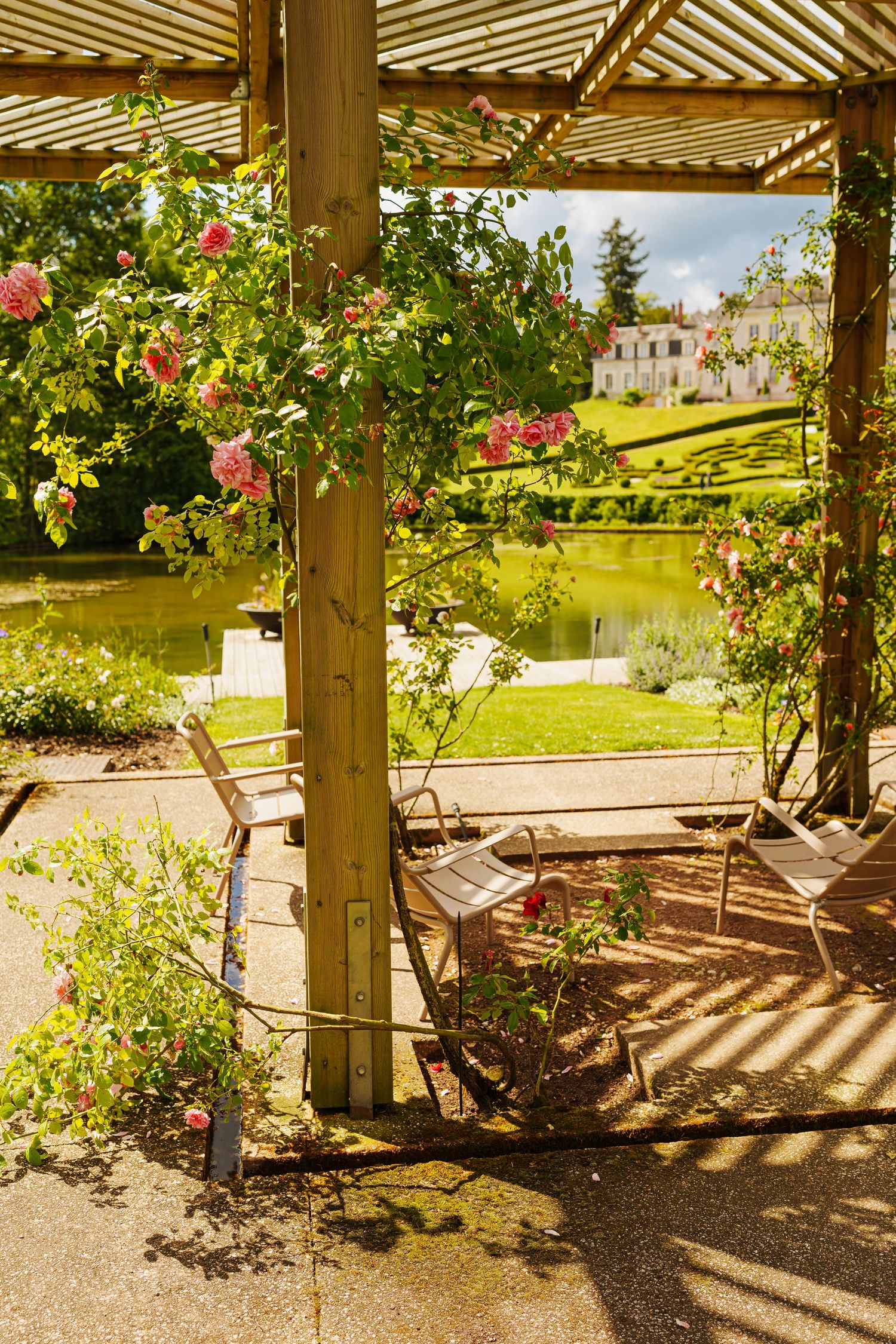 Charming garden pergola with pink roses, wooden chairs, and a serene pond view near a classic French chateau.
