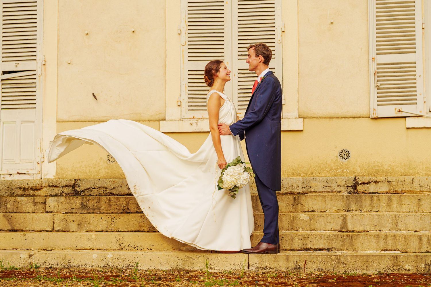 A romantic wedding portrait with a flowing white dress against a vintage building with shuttered windows.