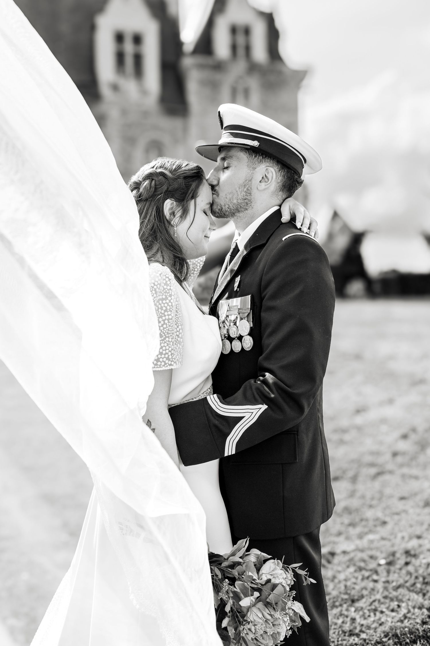 Black and white photo of a bride and groom kissing, groom in military uniform with medals, veil flowing in the wind.