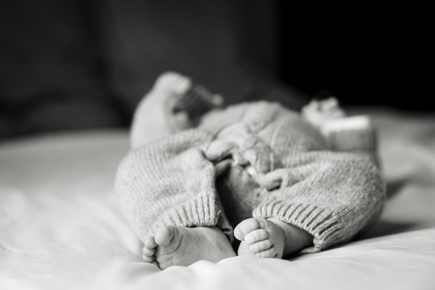 Black and white photo of newborn twins feet and legs in knit onesies lying on white blanket.