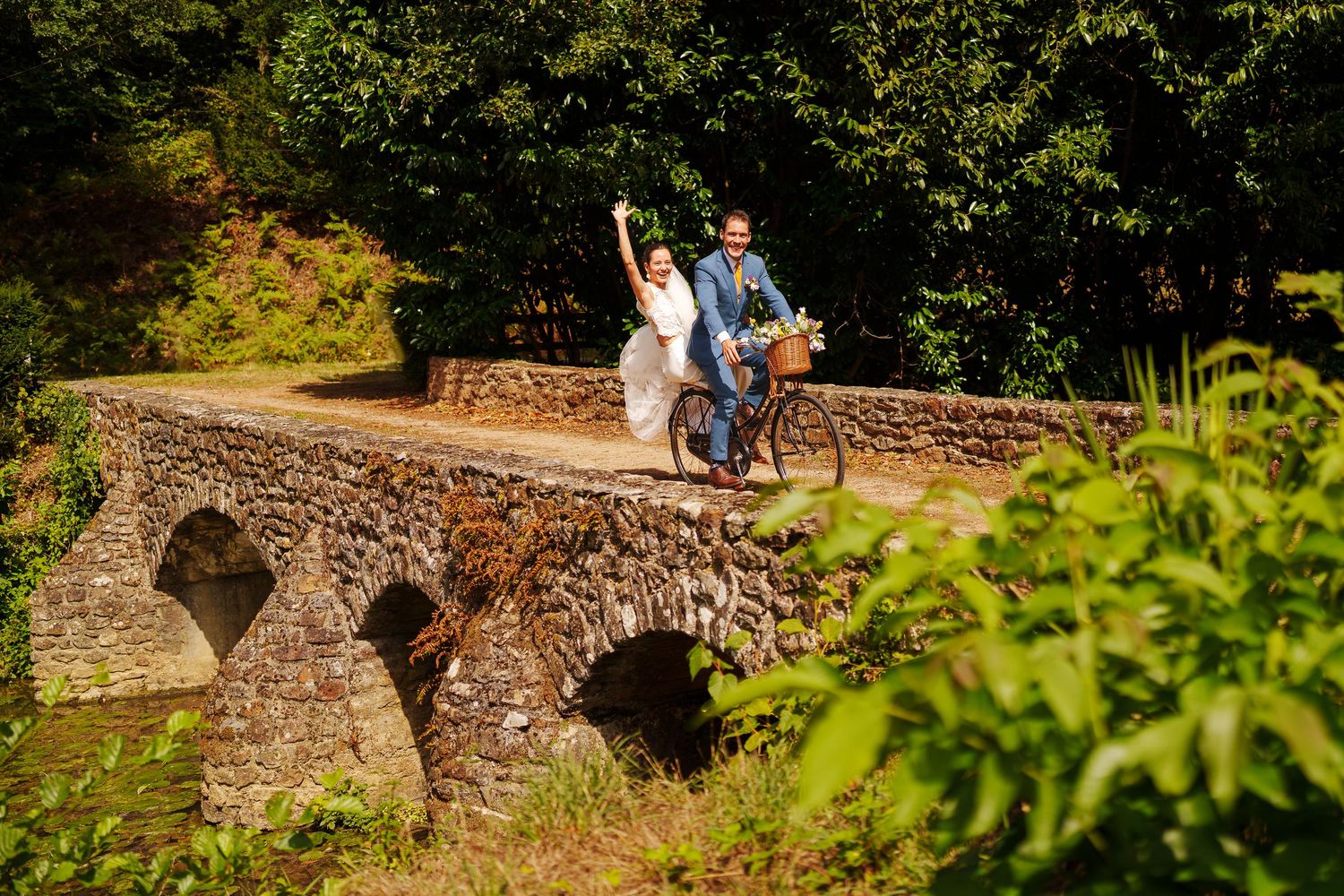 A cyclist crosses a historic stone arch bridge surrounded by lush green foliage on a sunny day.