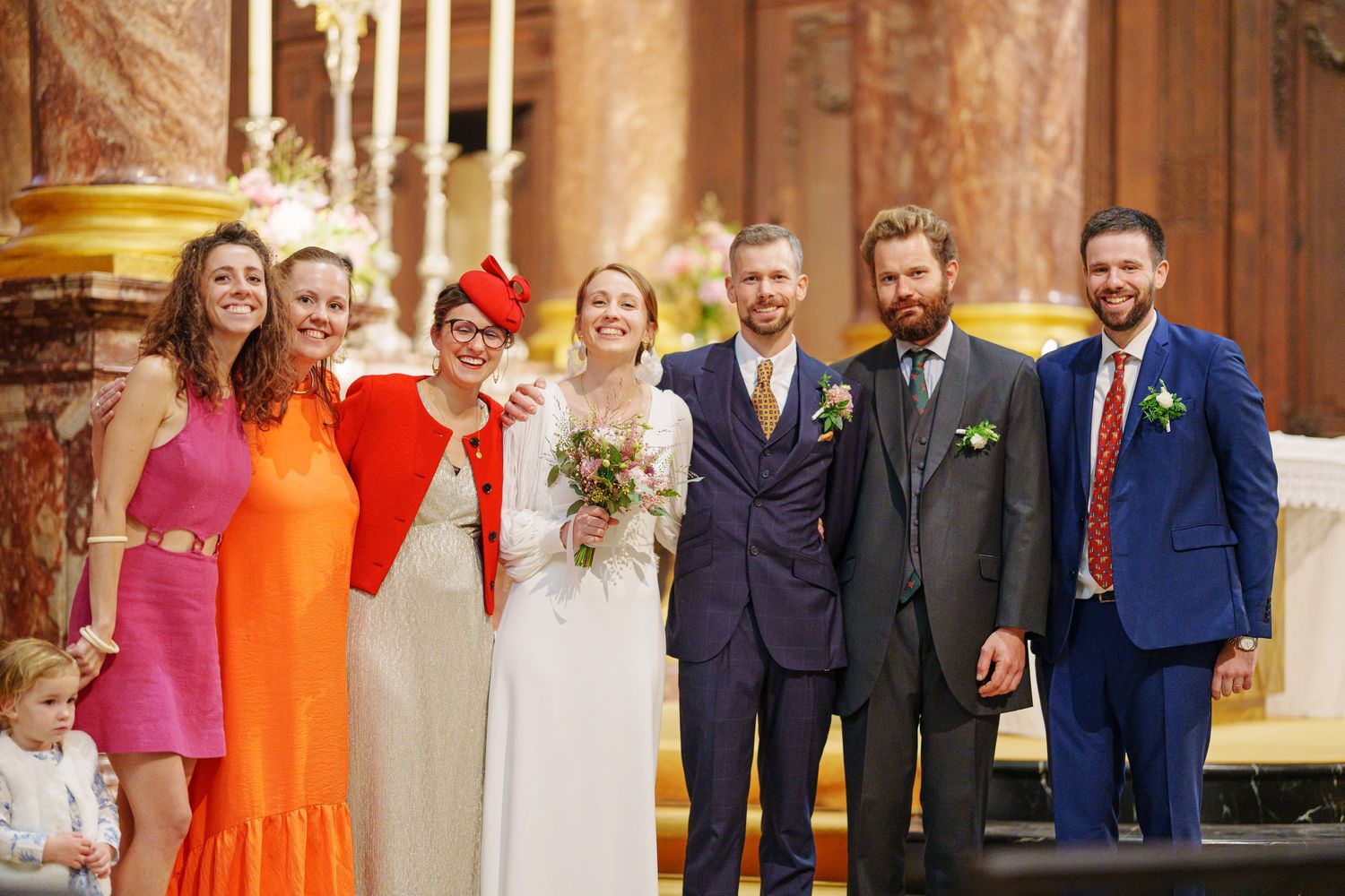 Wedding party members in colorful orange dresses and navy suits pose together at formal celebration.