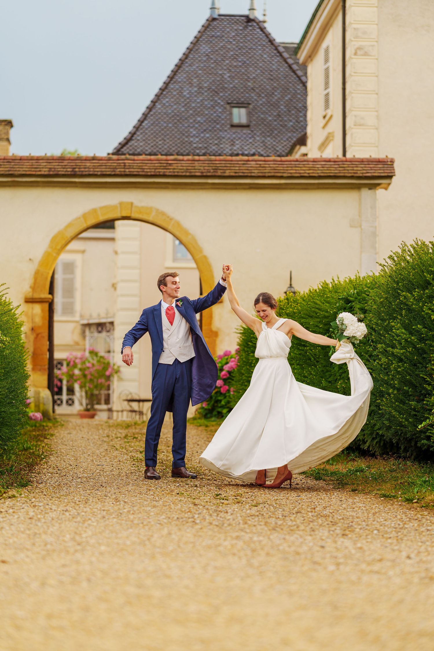 A couple in wedding attire dances and twirls together in front of a historic stone building on a gravel courtyard.
