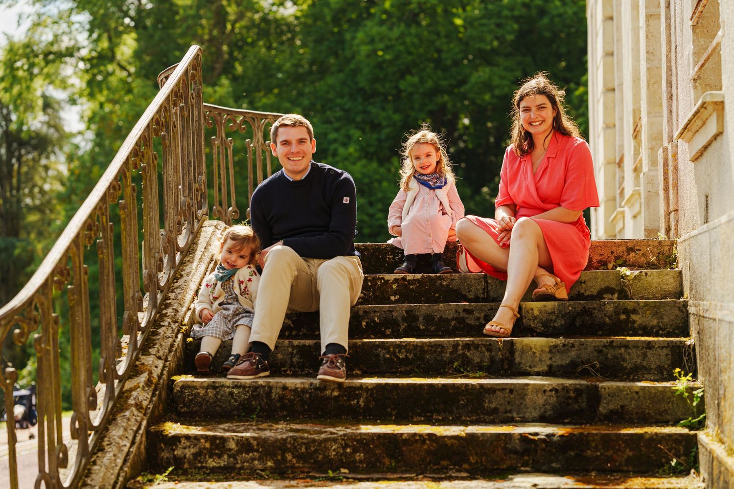 Family of four sitting on stone steps outdoors, with lush green trees in the background on a sunny day.