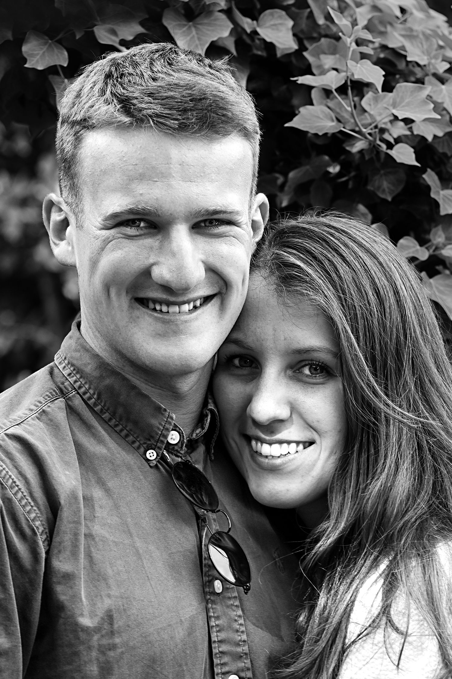 A black and white portrait of a couple smiling against foliage.