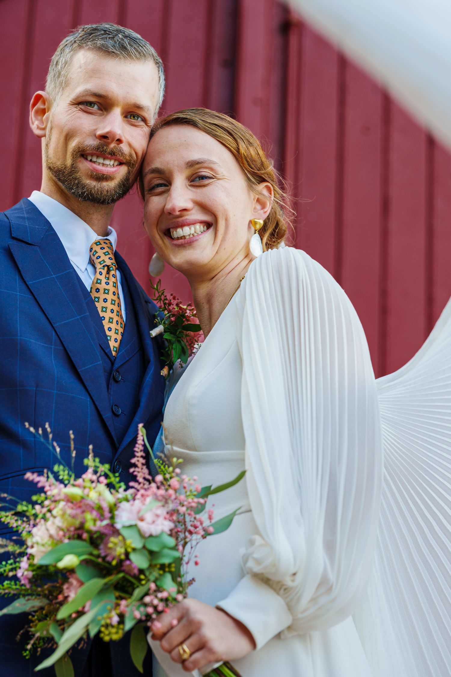 A joyful wedding couple embraces against a red barn wall while holding a bouquet of pink and purple wildflowers.