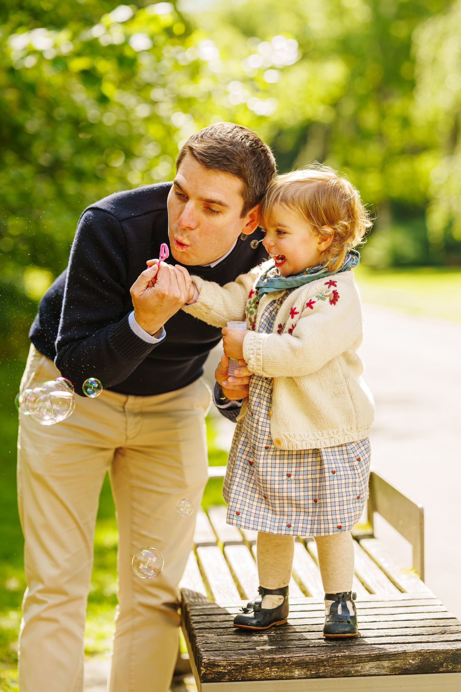 Father blowing soap bubbles with toddler daughter standing on park bench on sunny day.