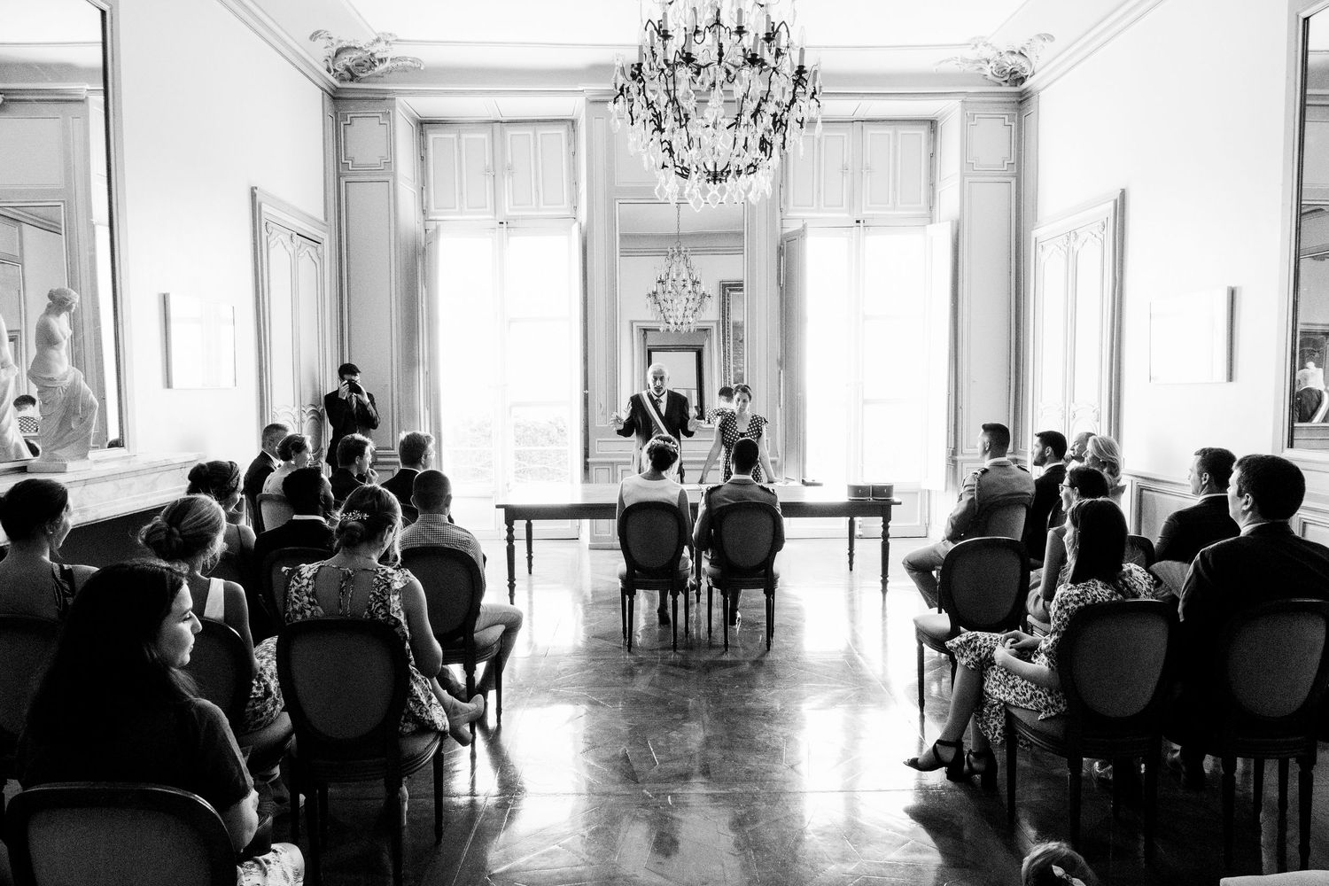 Wide panoramic view of an elegant white ballroom with chandelier and wedding guests.