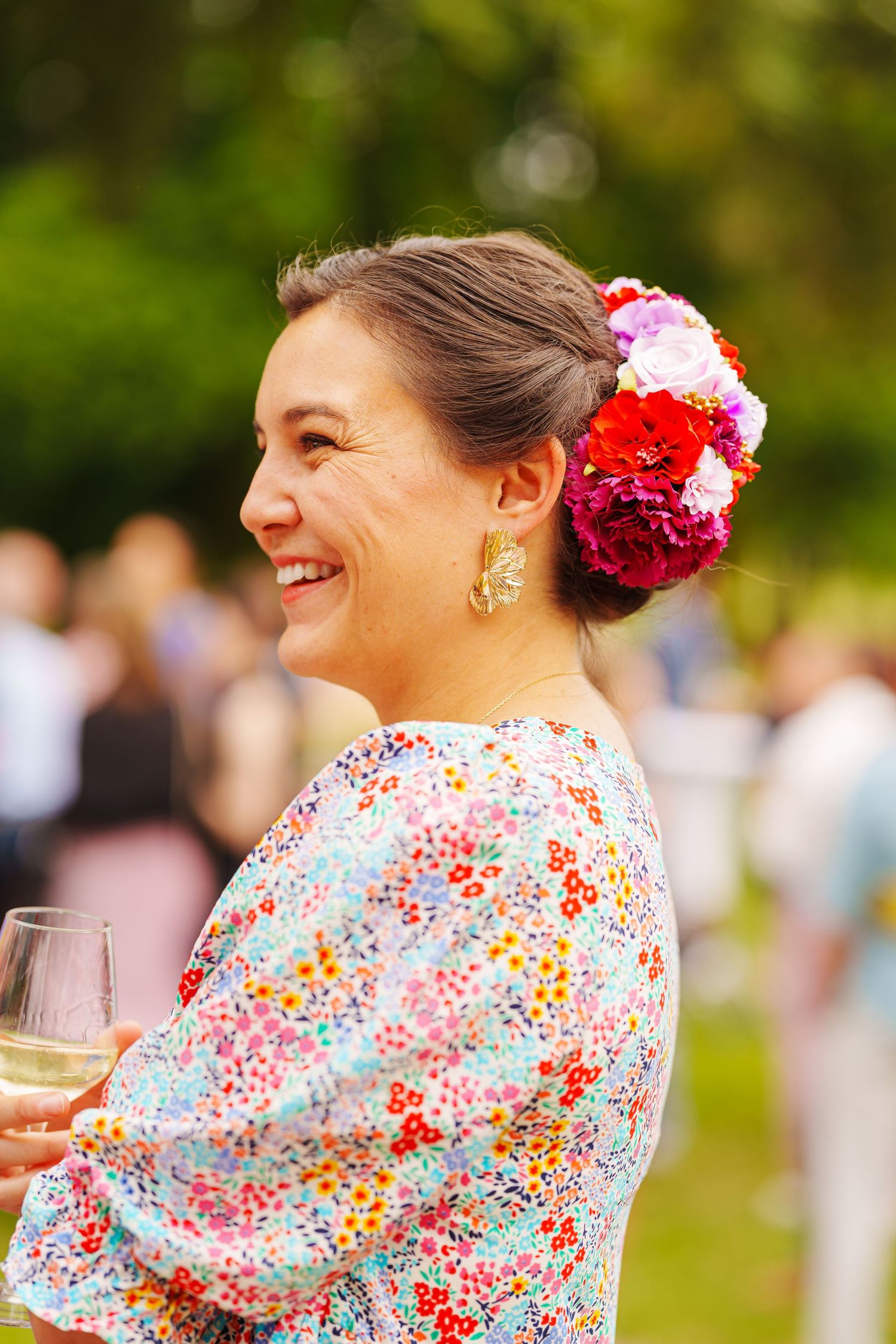 A woman in a floral dress smiles while wearing bright flowers in her hair at an outdoor event.