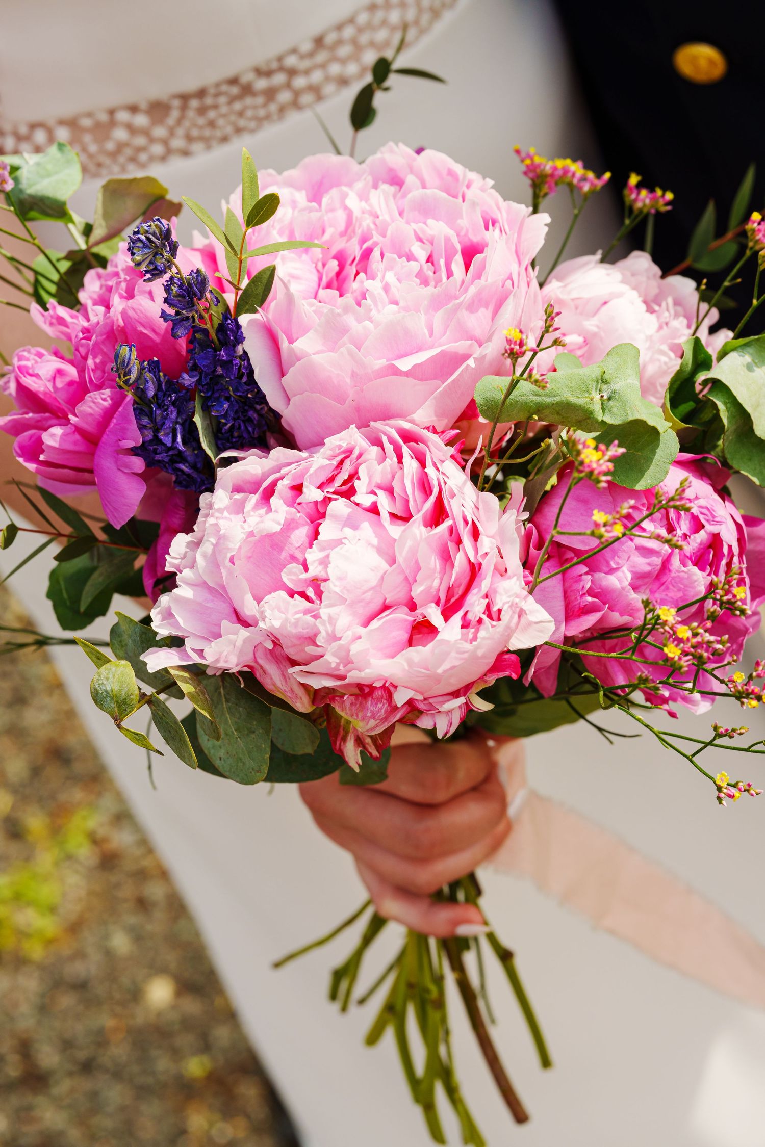 Hand holding a bridal bouquet of pink peonies, purple hyacinths, and greenery at a wedding.