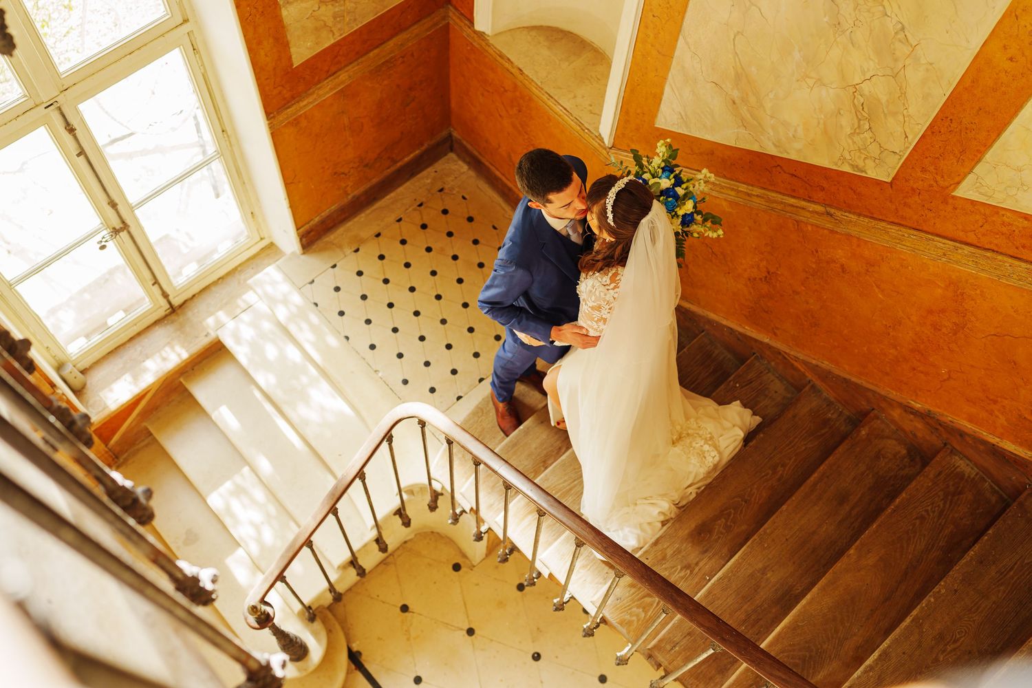Two people embrace on a winding staircase with tiled floors and orange walls in an elegant building interior.