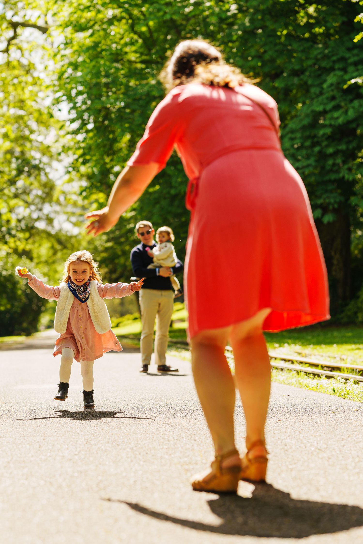 A joyful little girl runs toward her mother in a sunny park while father holds a baby in the background.