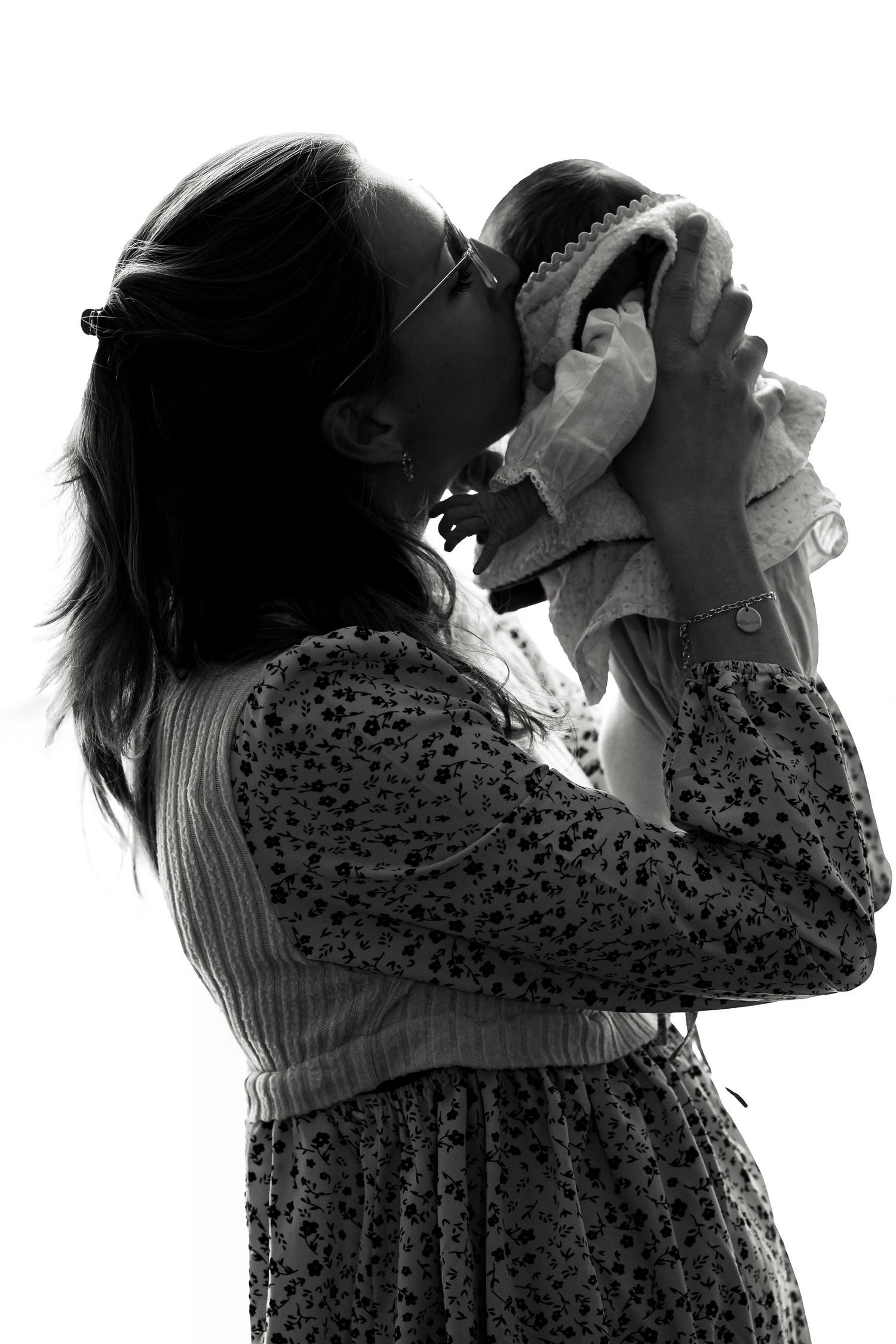 Black and white silhouette of a mother lifting and kissing her baby, wearing a floral dress and sunglasses.