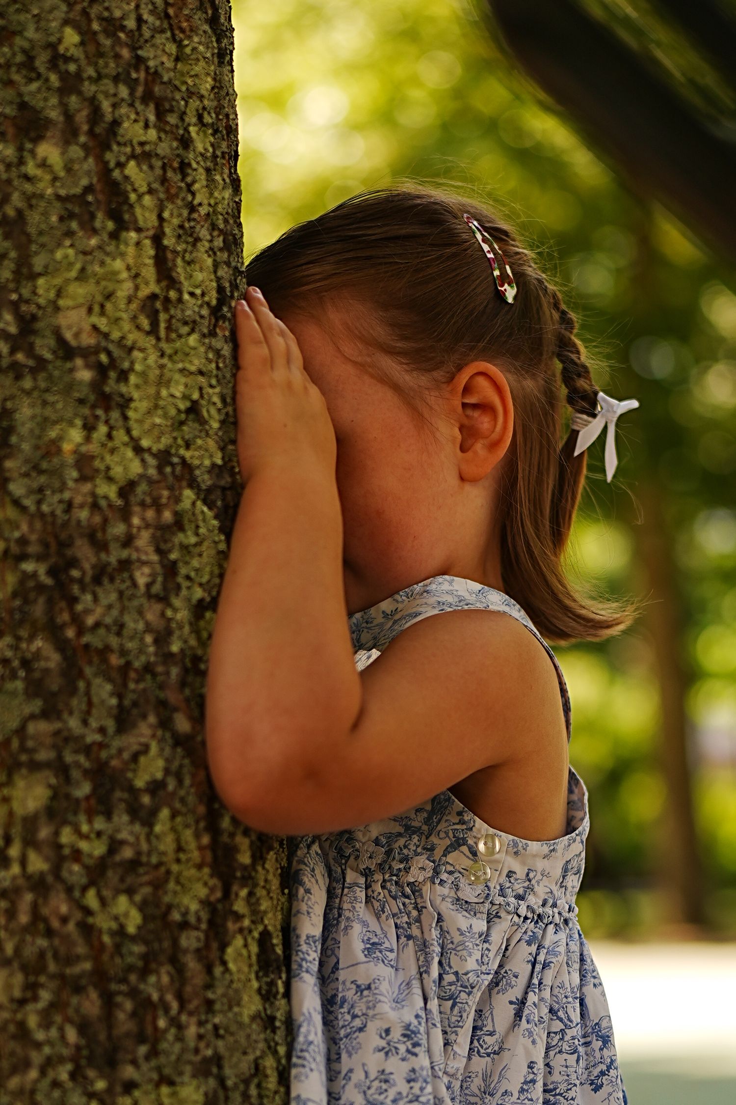 Child in floral dress playing hide and seek behind a large tree trunk.