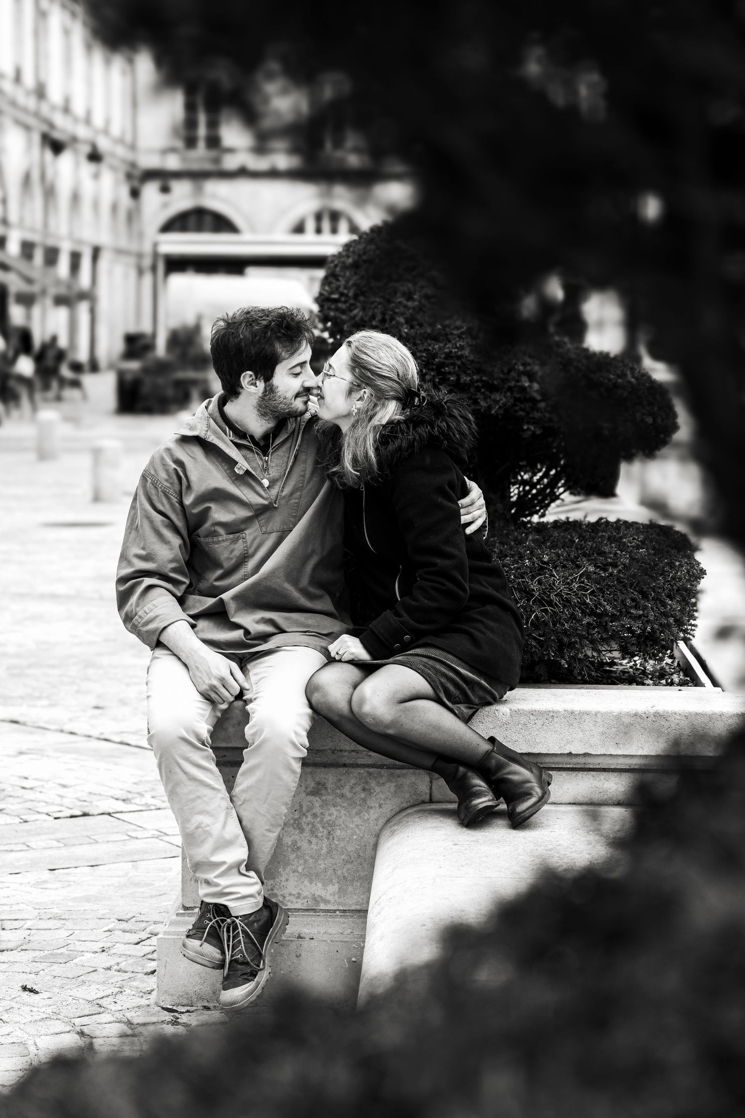 Black and white photo of a romantic couple kissing on a stone bench in a European city plaza.