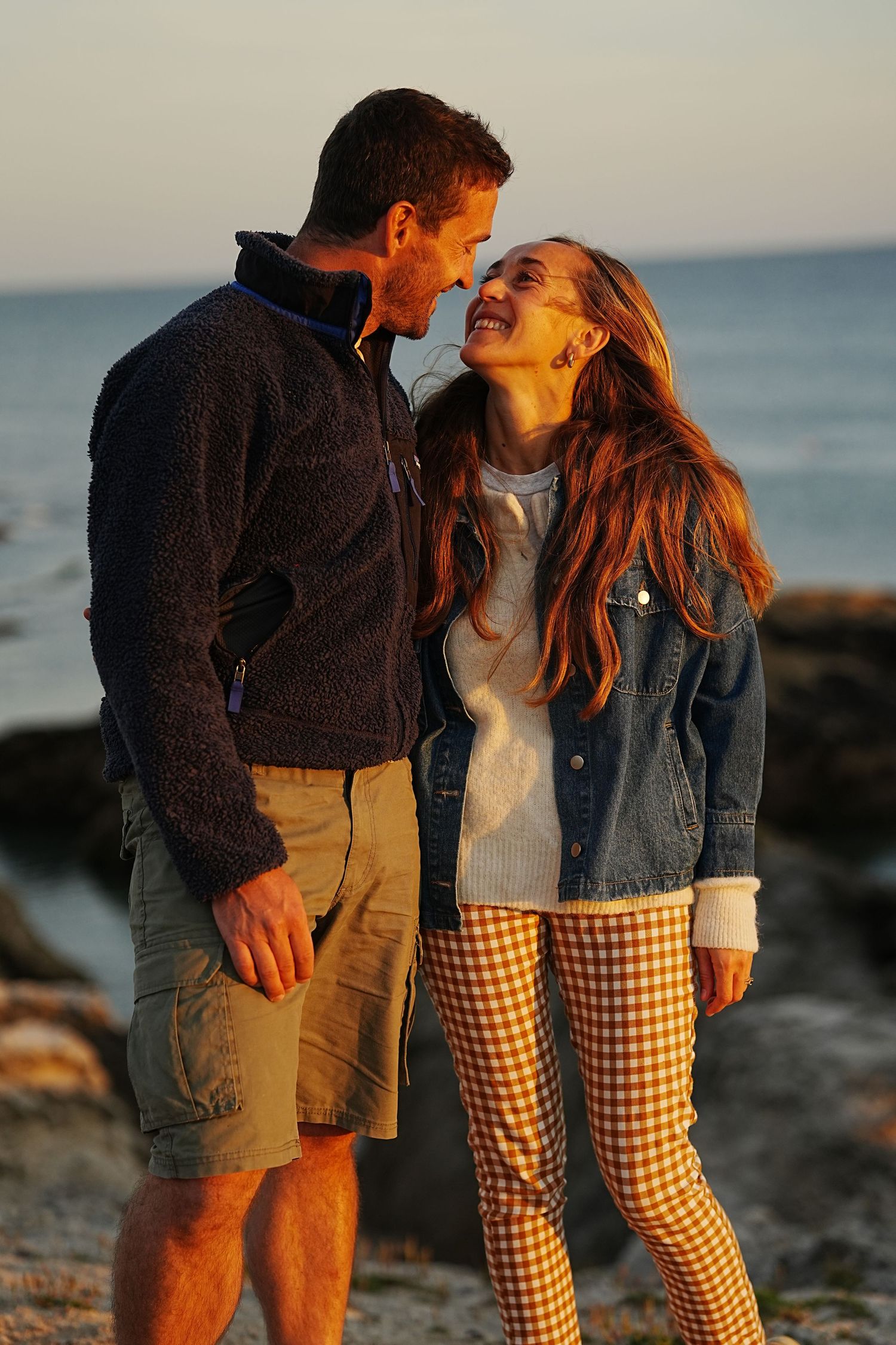 Couple sharing a romantic moment at golden hour on a rocky beach, woman in plaid pants and denim jacket.