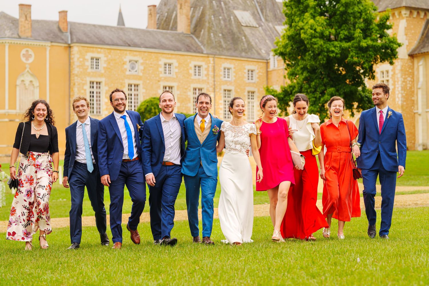 Wedding party members walk and pose together on a green lawn in front of a historic stone building.