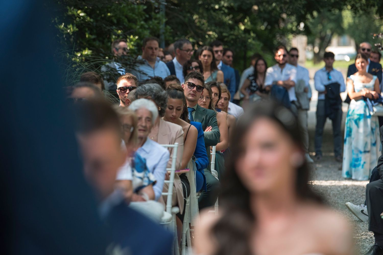 Wedding guests seated outdoors in shaded garden aisle watch ceremony unfold on sunny day.