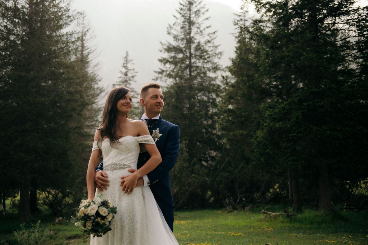 Bride and groom embracing in a lush forest setting, with tall evergreen trees in the misty background.